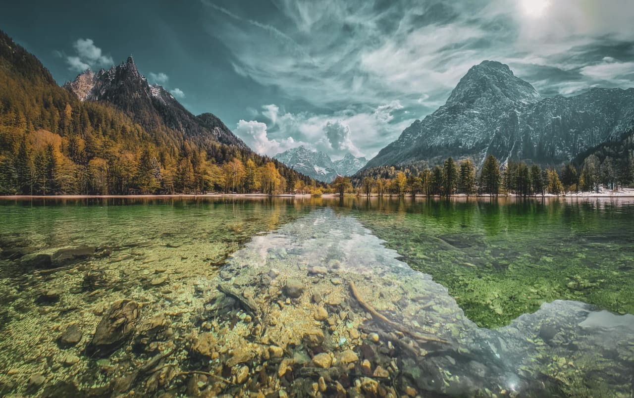 A clear, tranquil lake reflects the majestic mountains that surround it. The surface of the water is shades of green and blue, while rocks and pebbles are visible in the shallow parts.