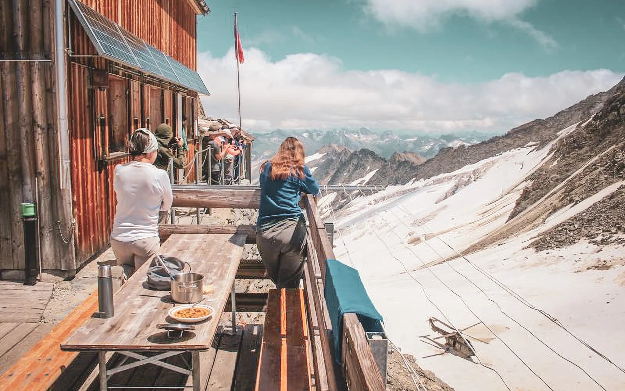 A wooden deck overlooks an impressive alpine landscape, where majestic mountains rise up under a partly cloudy sky. On the left, the wooden Oberaarjochhutte with solar panels and a flying flag.