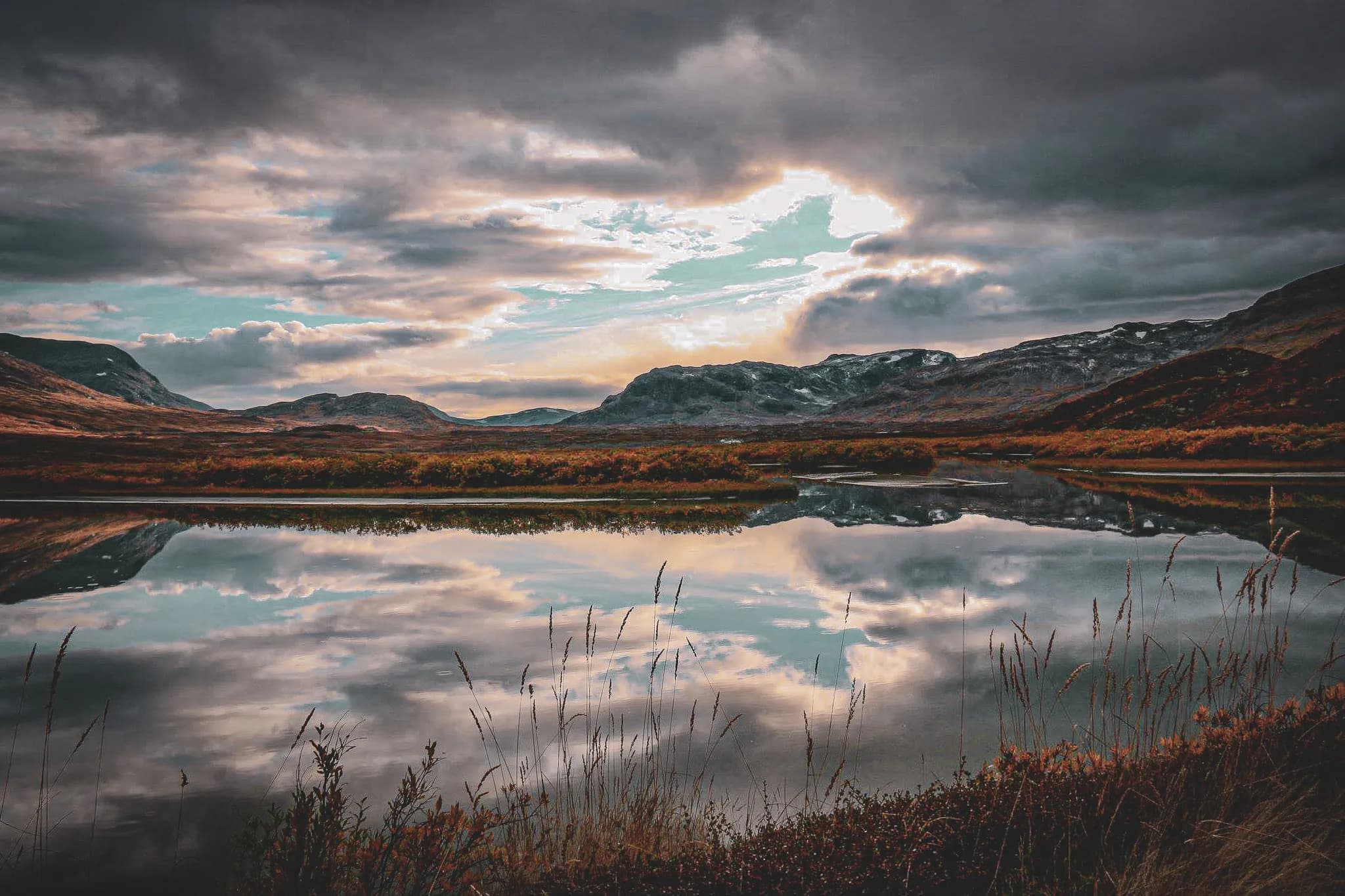 A mountainous landscape at dusk, with a calm lake reflecting the clouds and surrounding hills. The mountains rise majestically in the background, while the warm hues of the