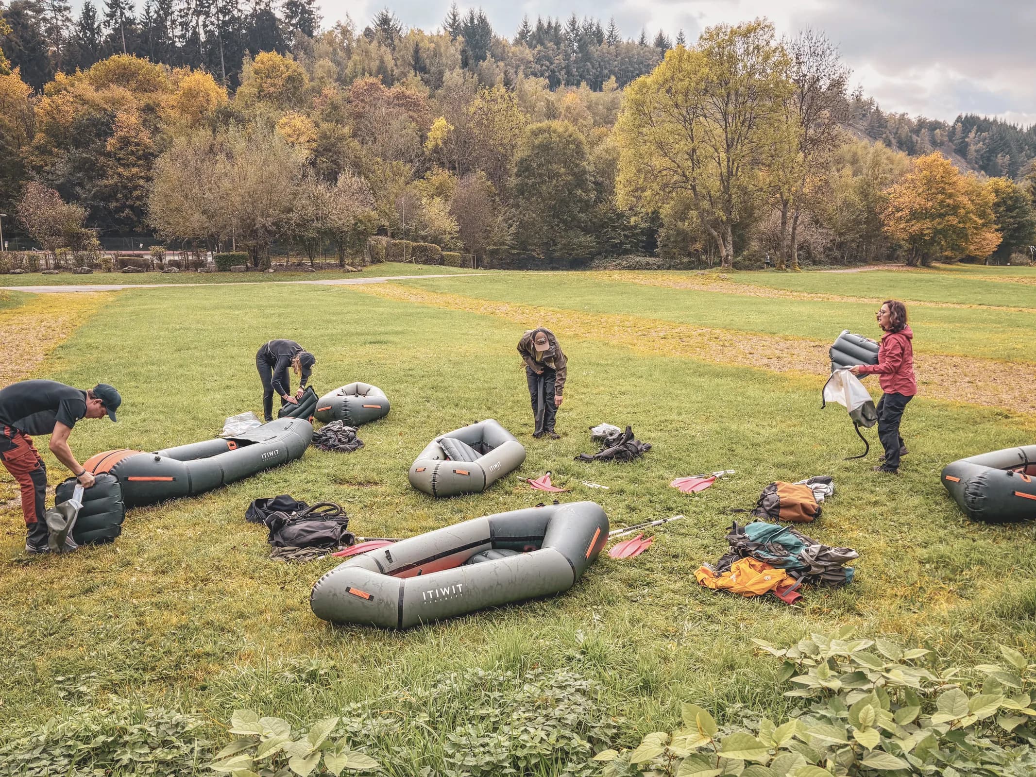 A group prepares packrafts on a green field surrounded by colourful autumn forests.