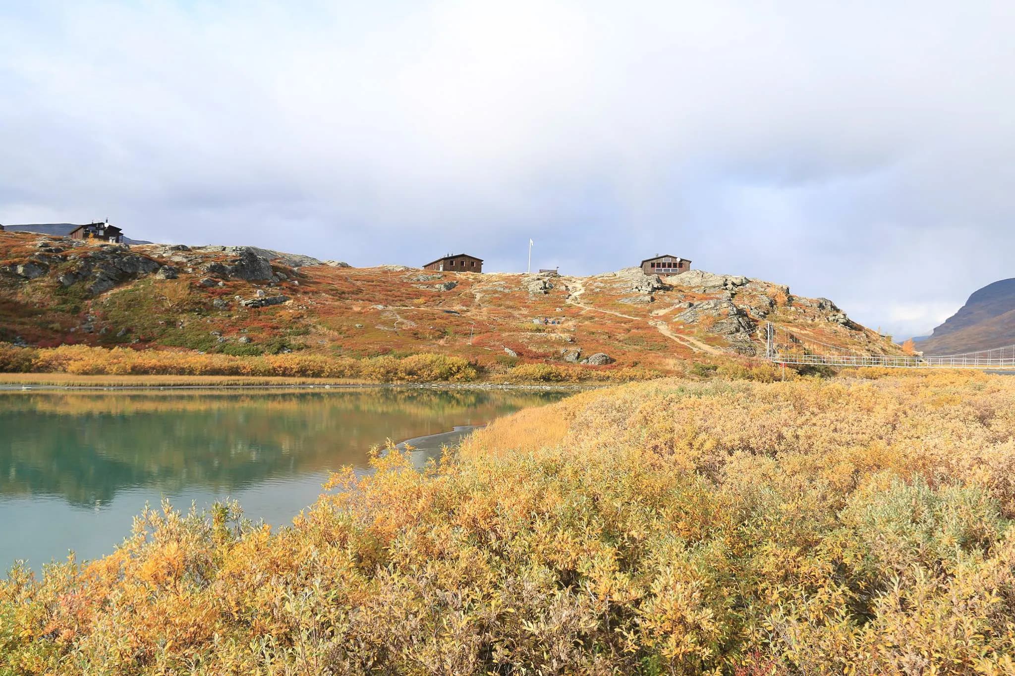 A peaceful Alpine landscape with a lake in the foreground, surrounded by golden and orange autumn foliage. In the background, wooden chalets perch on a rocky hill,
