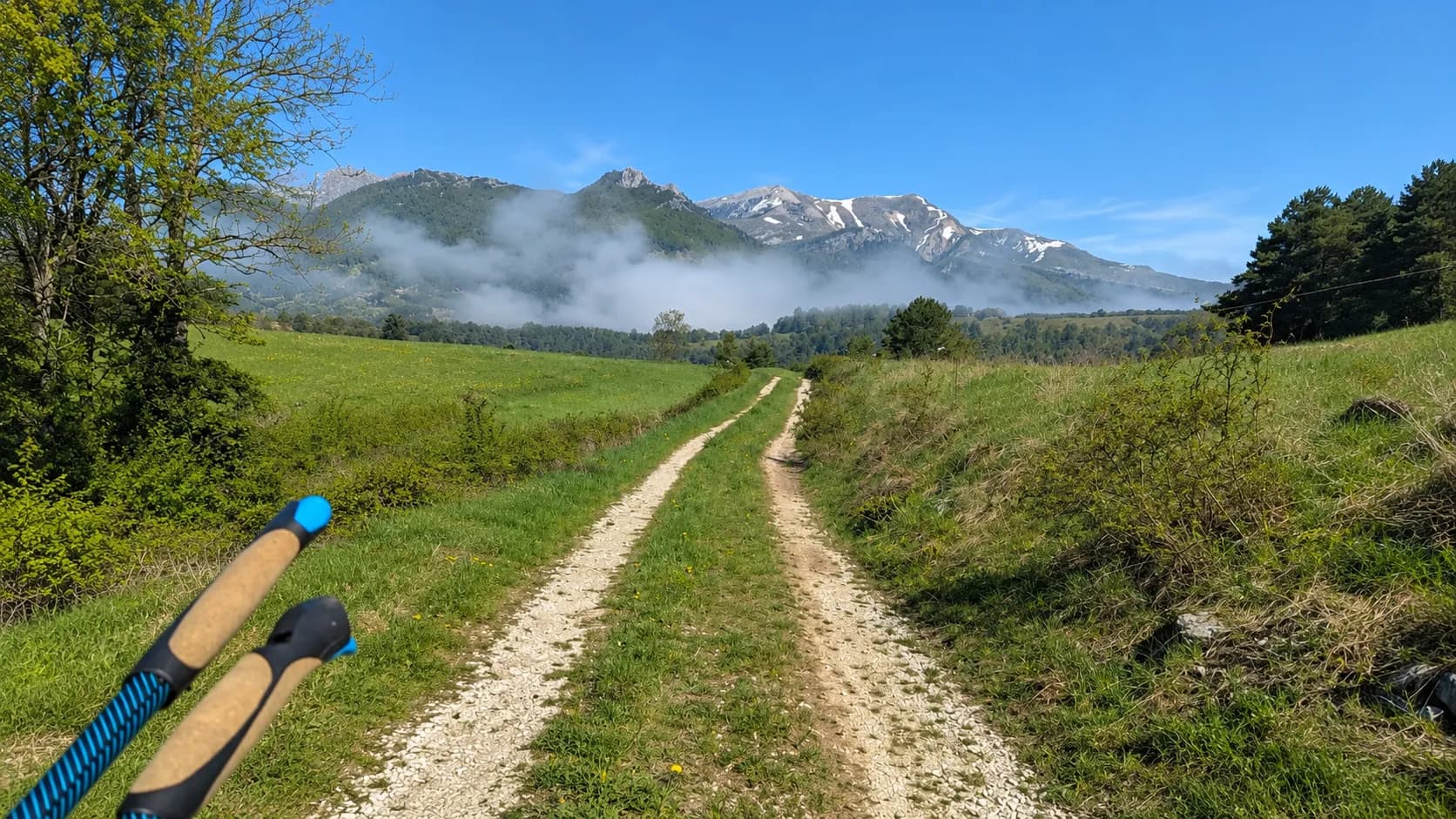 Chemin de gravier verdoyant avec montagnes en arrière-plan, ciel bleu et nuages légers.