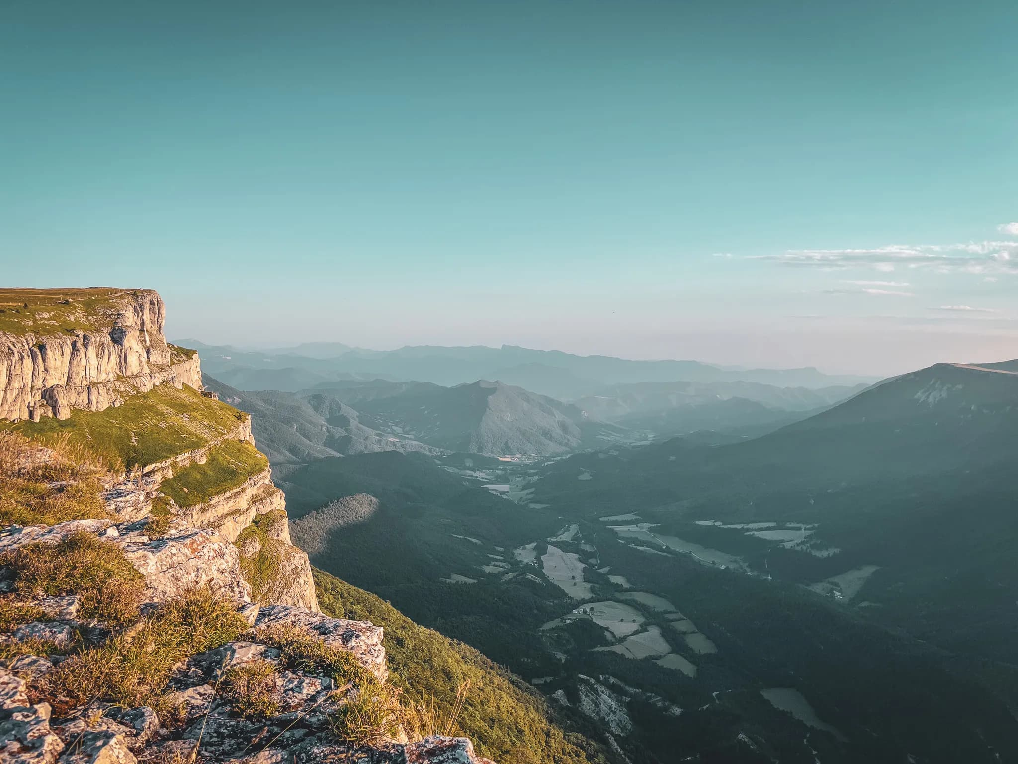 A mountainous panorama stretches out beneath a clear sky, with layers of rock overlooking lush green valleys. Peaks rise up in the background, creating an impressive perspective, while
