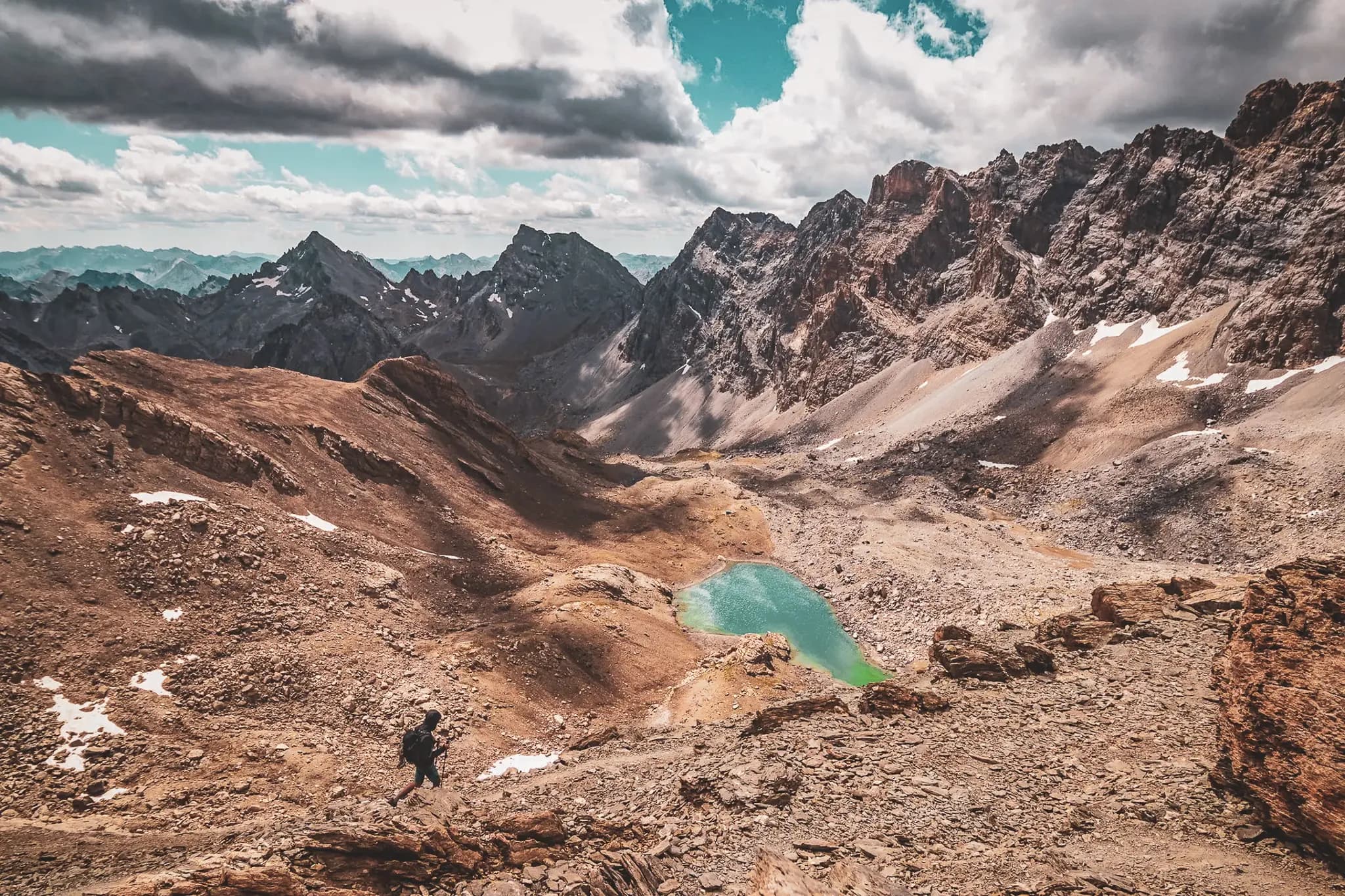 A mountainous landscape with steep, rocky peaks under a partly cloudy sky. In the foreground, a lone hiker can be seen climbing the rugged terrain. In the background, a small lake with