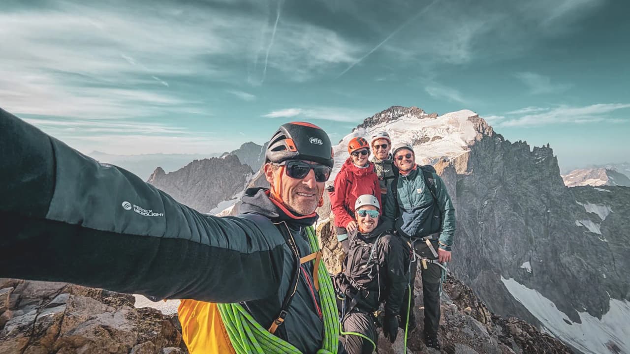 Groupe d'alpinistes souriants au sommet, entouré de montagnes majestueuses et de neige.
