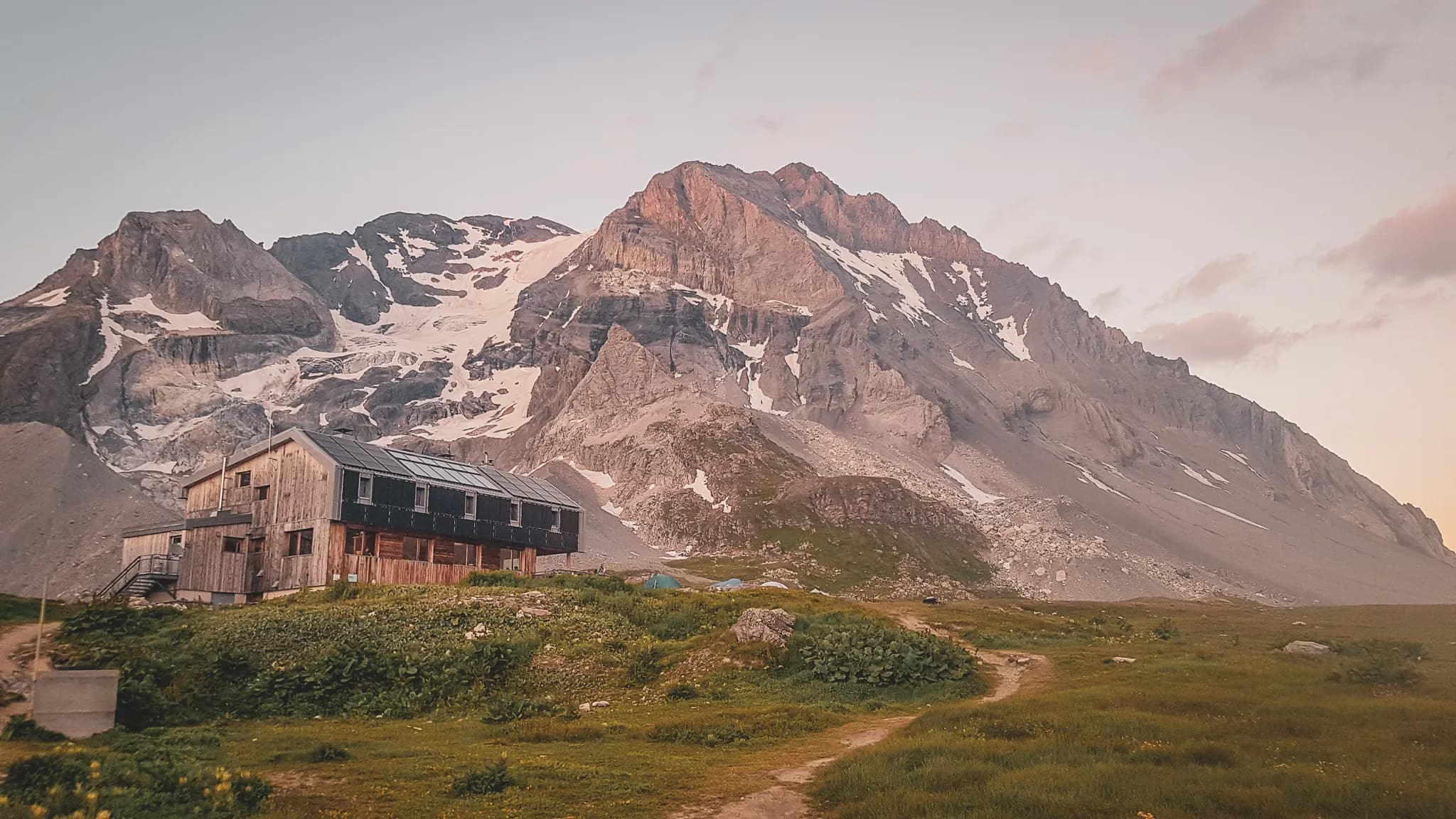 Un chalet en bois est situé au pied d'une montagne majestueuse, entouré de paysages naturels. La montagne, avec ses sommets escarpés et ses zones de glace résiduelle, se dresse fièrement en arrière-plan. Le ciel