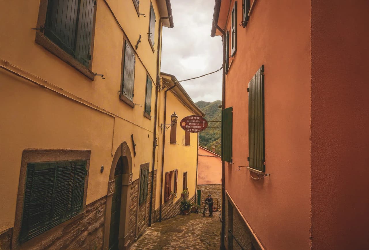 An alleyway lined with yellow and red houses in a small village in Southern Europe. A cyclist is standing in the middle of the lane.