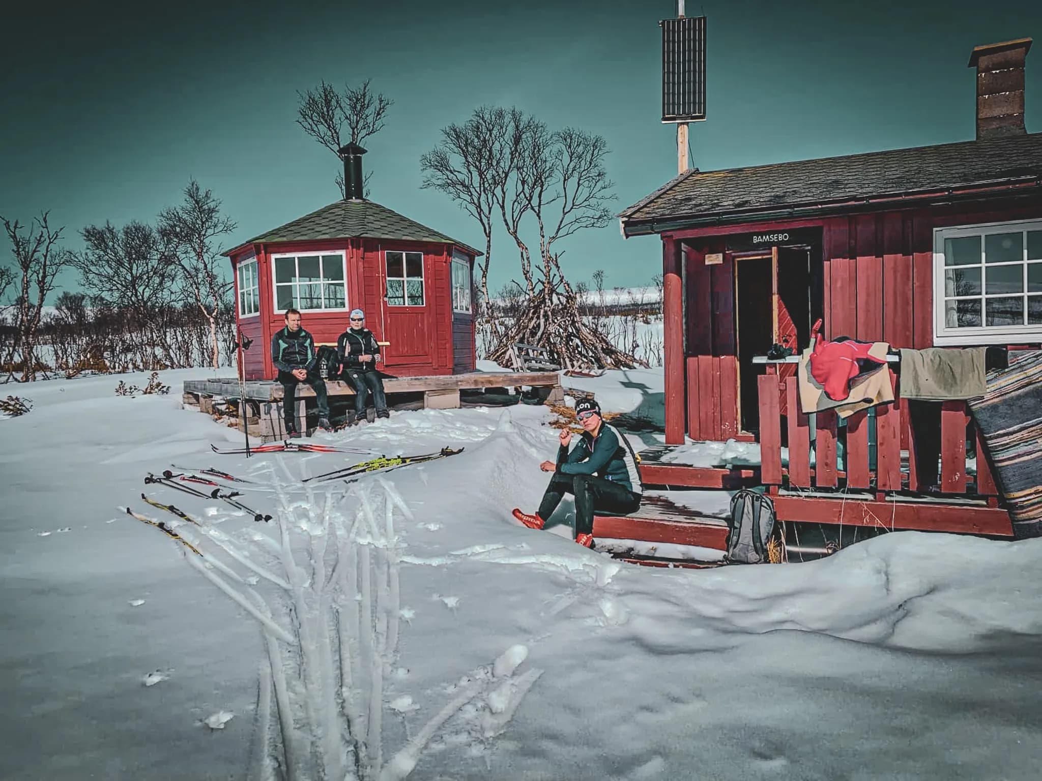 A red wooden chalet stands in the middle of a snowy landscape, surrounded by bare trees. Two people, dressed in sporty outfits, are sitting on the terrace, soaking up the sun. Nearby