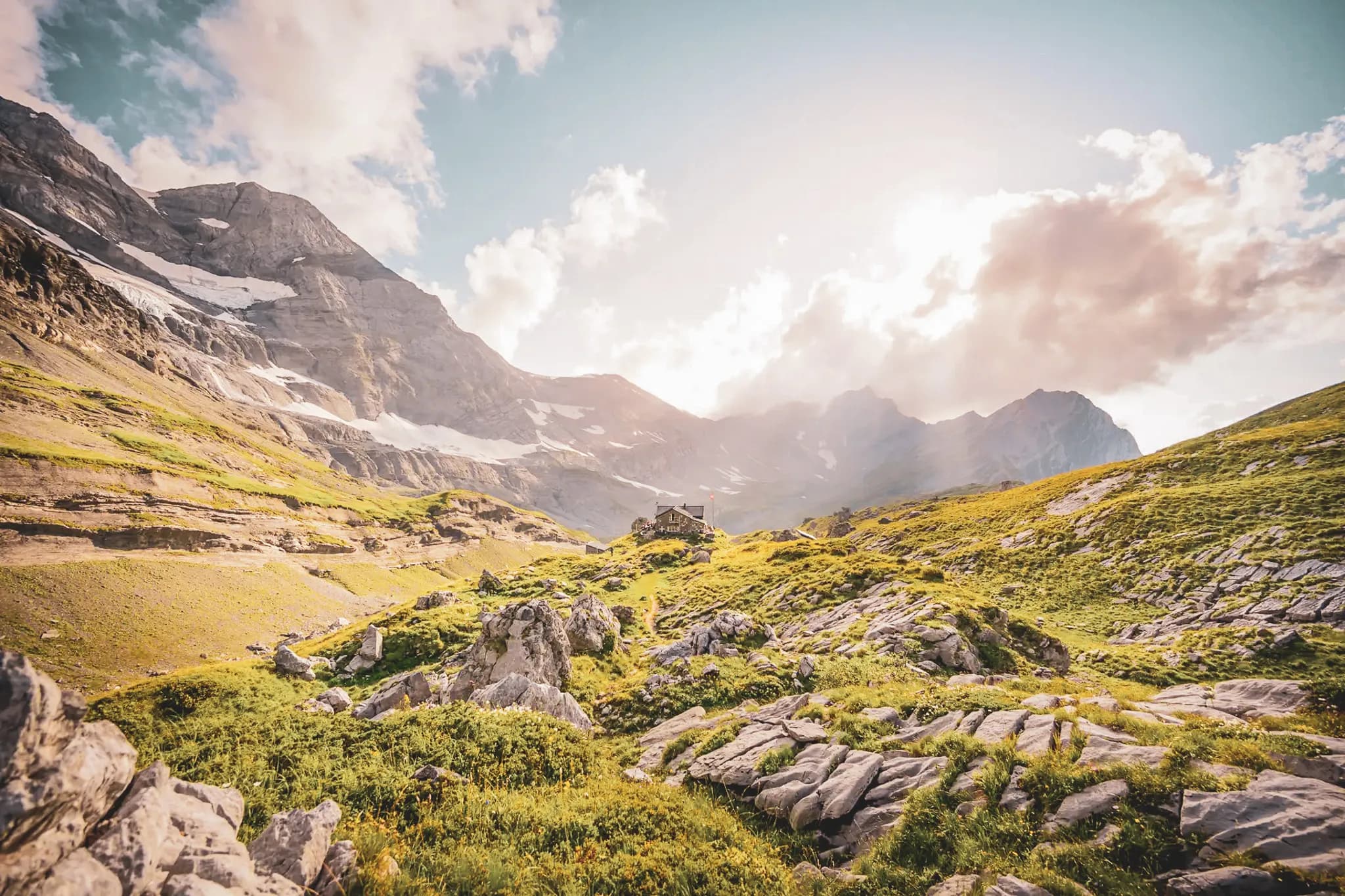 A spectacular mountain landscape of green slopes and rocks, bathed in golden light. In the centre, a charming wooden structure, probably a chalet or refuge, stands on a meadow.