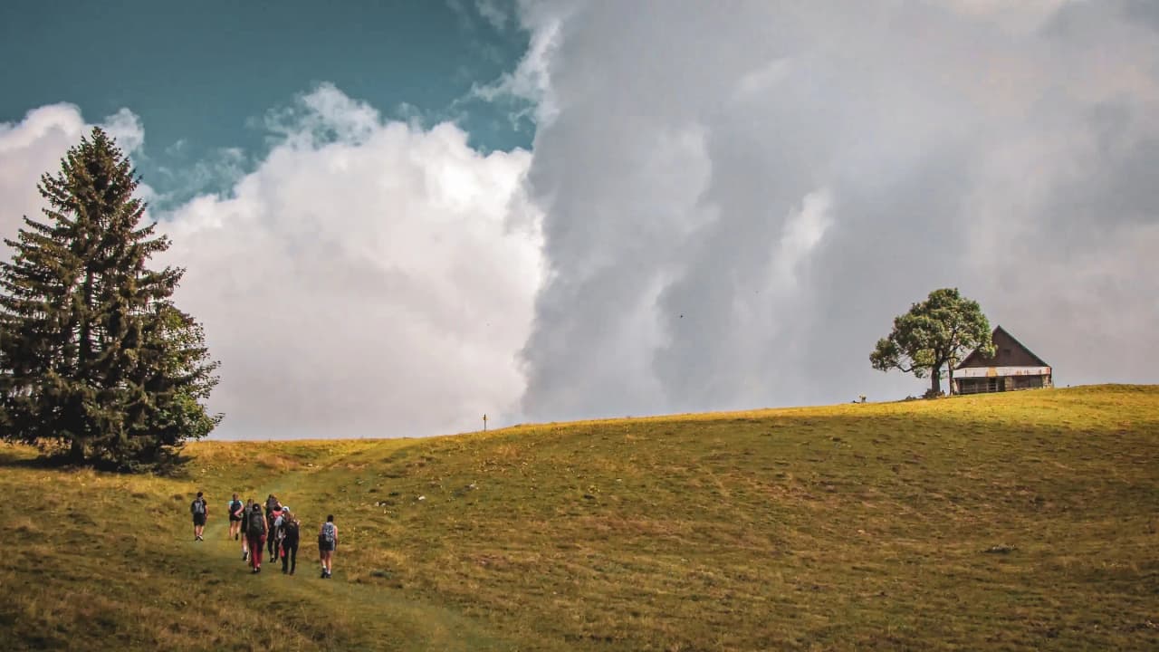 Un groupe de randonneurs sur une prairie verdoyante, avec un chalet et des nuages majestueux.