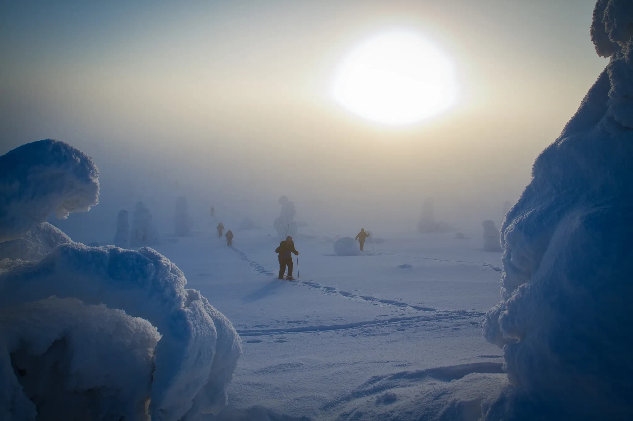 A snowy landscape shrouded in a light mist, where silhouettes of adventurers move through the snow. Majestic ice formations rise up on either side, while a pale sun emerges from the horizon.