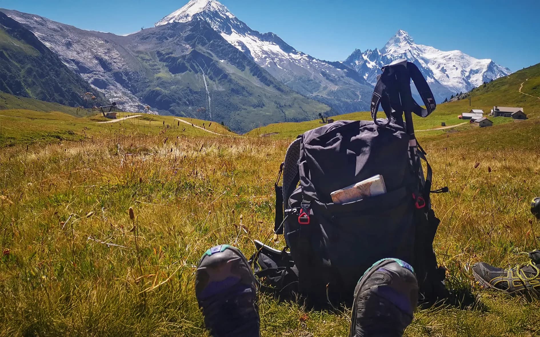 A sunny Alpine landscape with majestic mountains and snow-capped peaks in the background. In the foreground, a black rucksack lies on the green grass, with a bottle of water and a map.