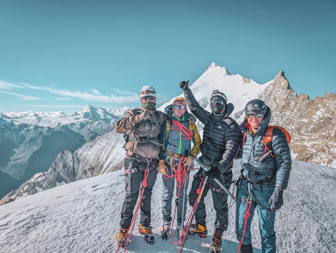 Four climbers on a snow-covered summit, smiling, with breathtaking mountain scenery in the background.