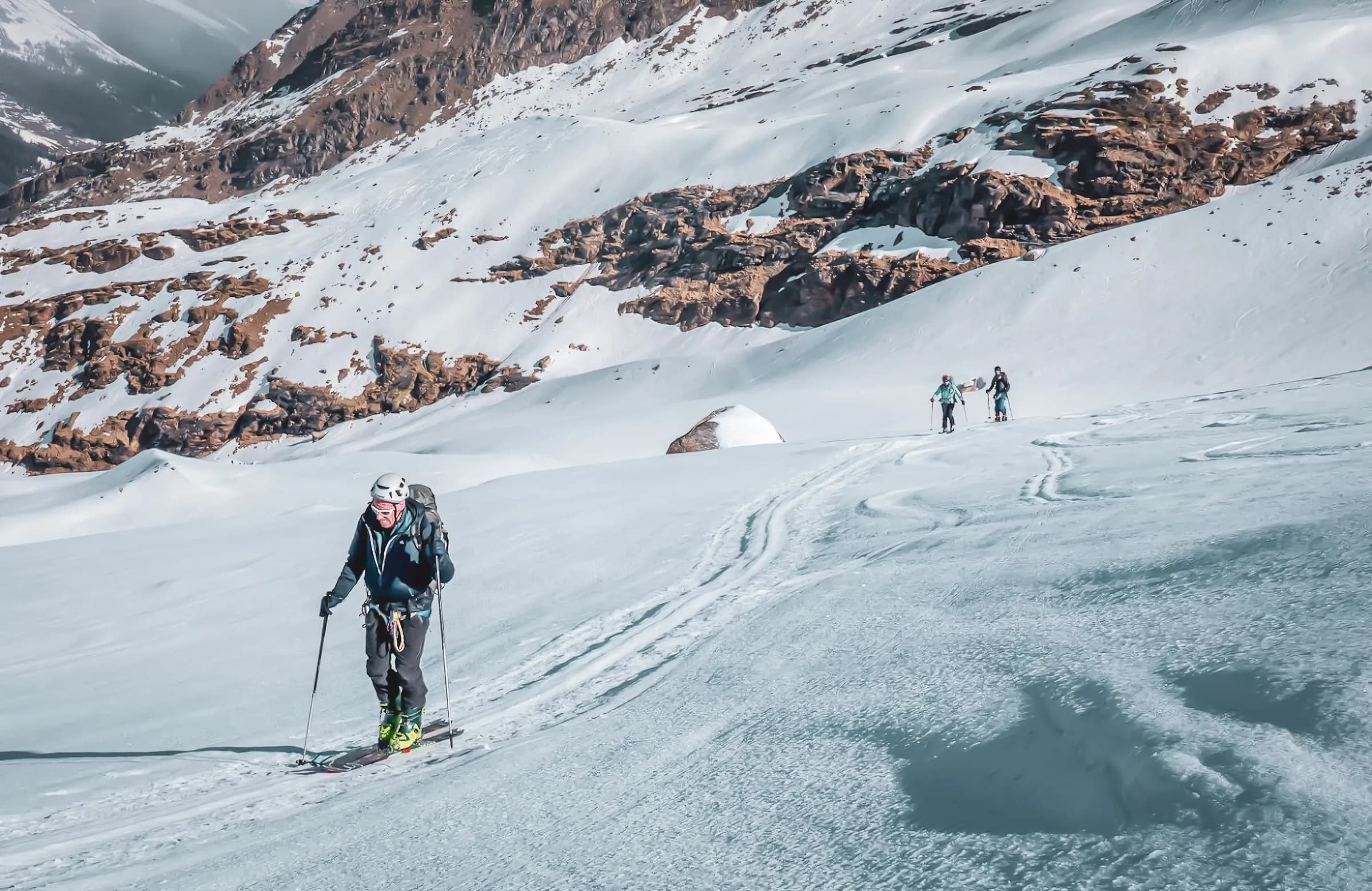 A skier progressing on a snow-covered surface, surrounded by impressive mountain scenery. The skier is equipped for mountaineering, with a rucksack and skis. In the background, rock formations