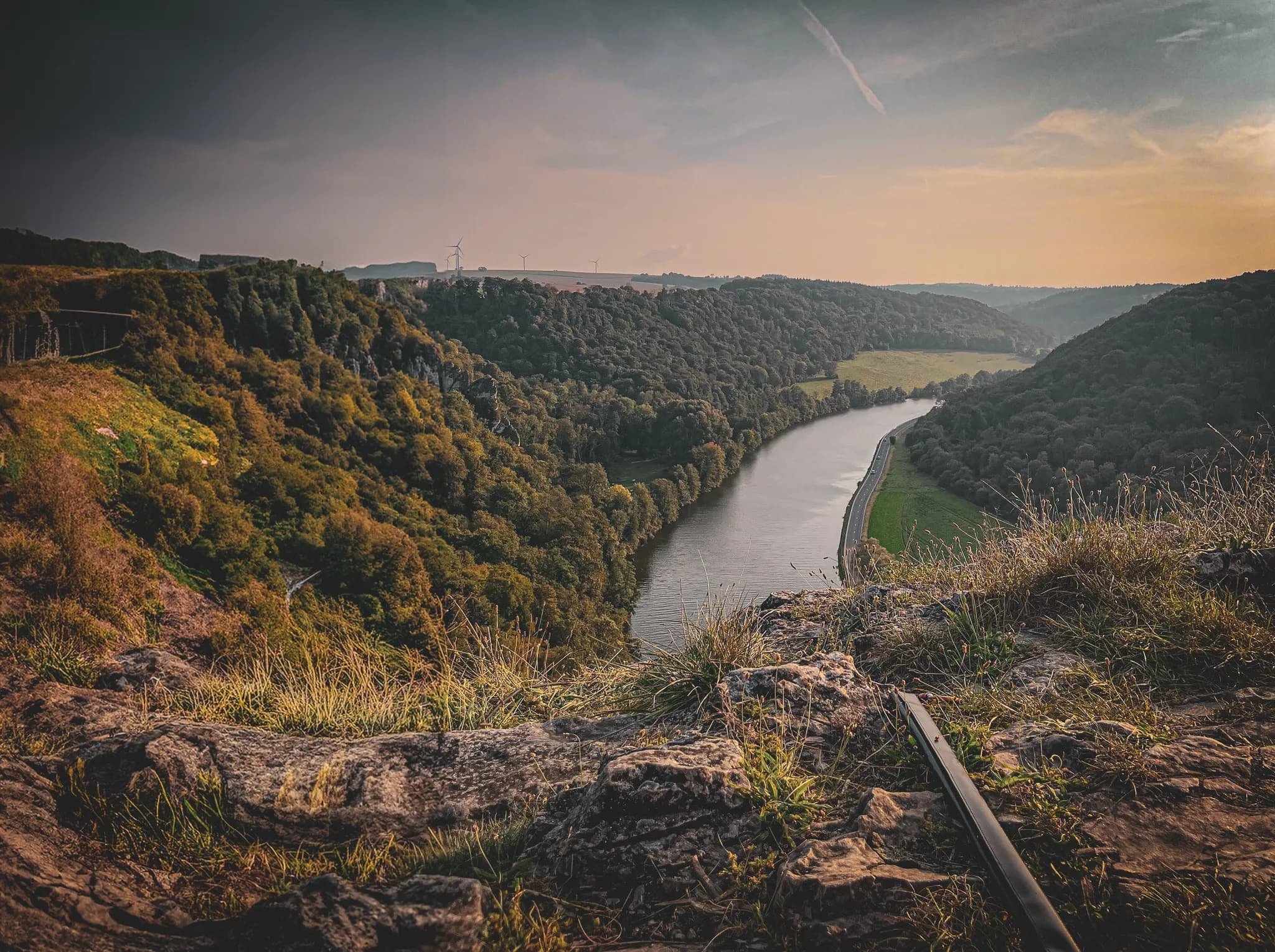 Een panorama van een groene vallei met in het midden een meanderende rivier. Beboste heuvels omzomen de oevers, terwijl een kronkelige weg langs een deel van de rivier loopt. De lucht is licht bewolkt, verlicht door de zon.