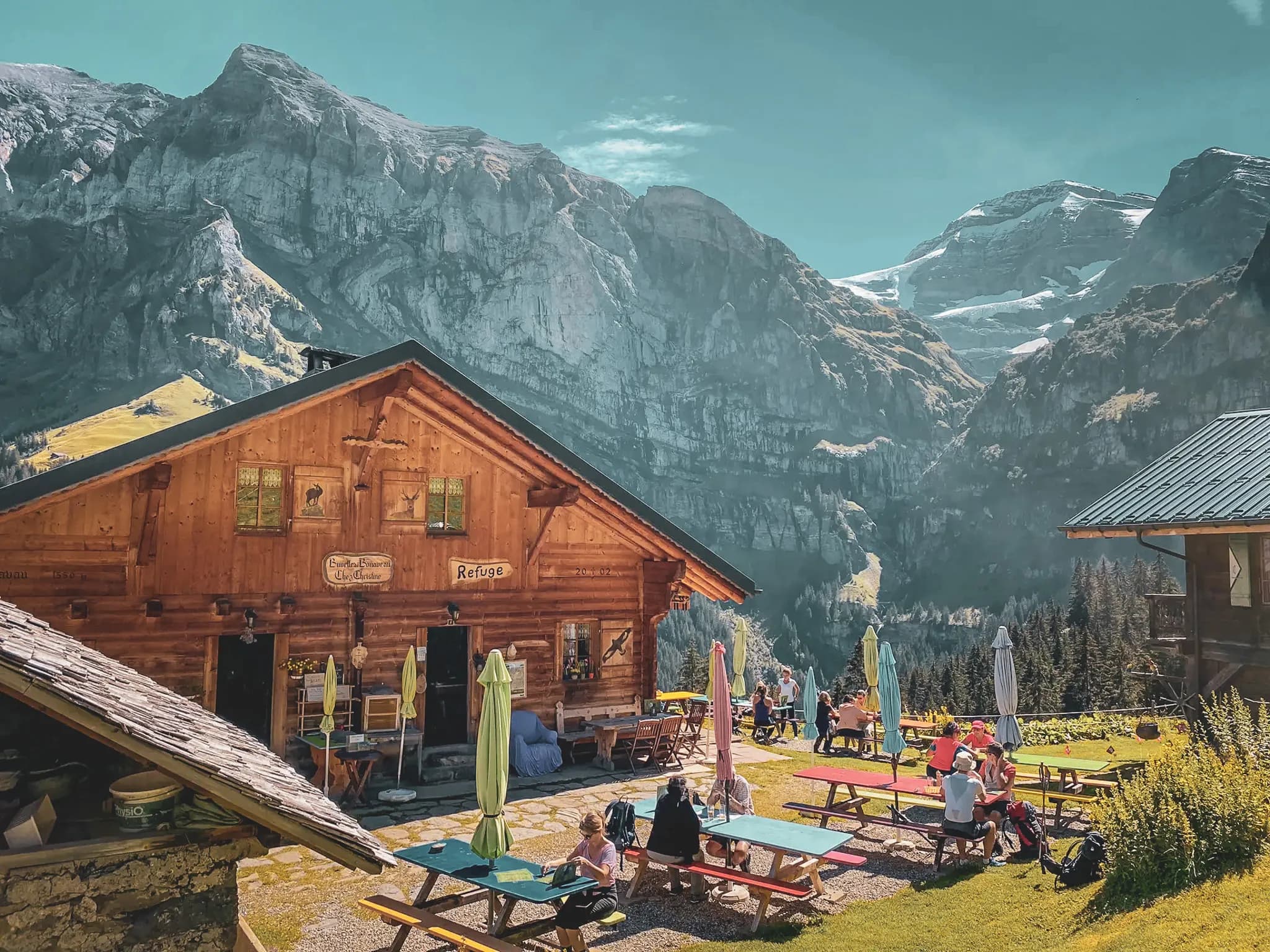A wooden hut, typical of the mountains, is surrounded by majestic mountains and greenery. Picnic tables adorned with colourful parasols welcome visitors enjoying the panoramic views.