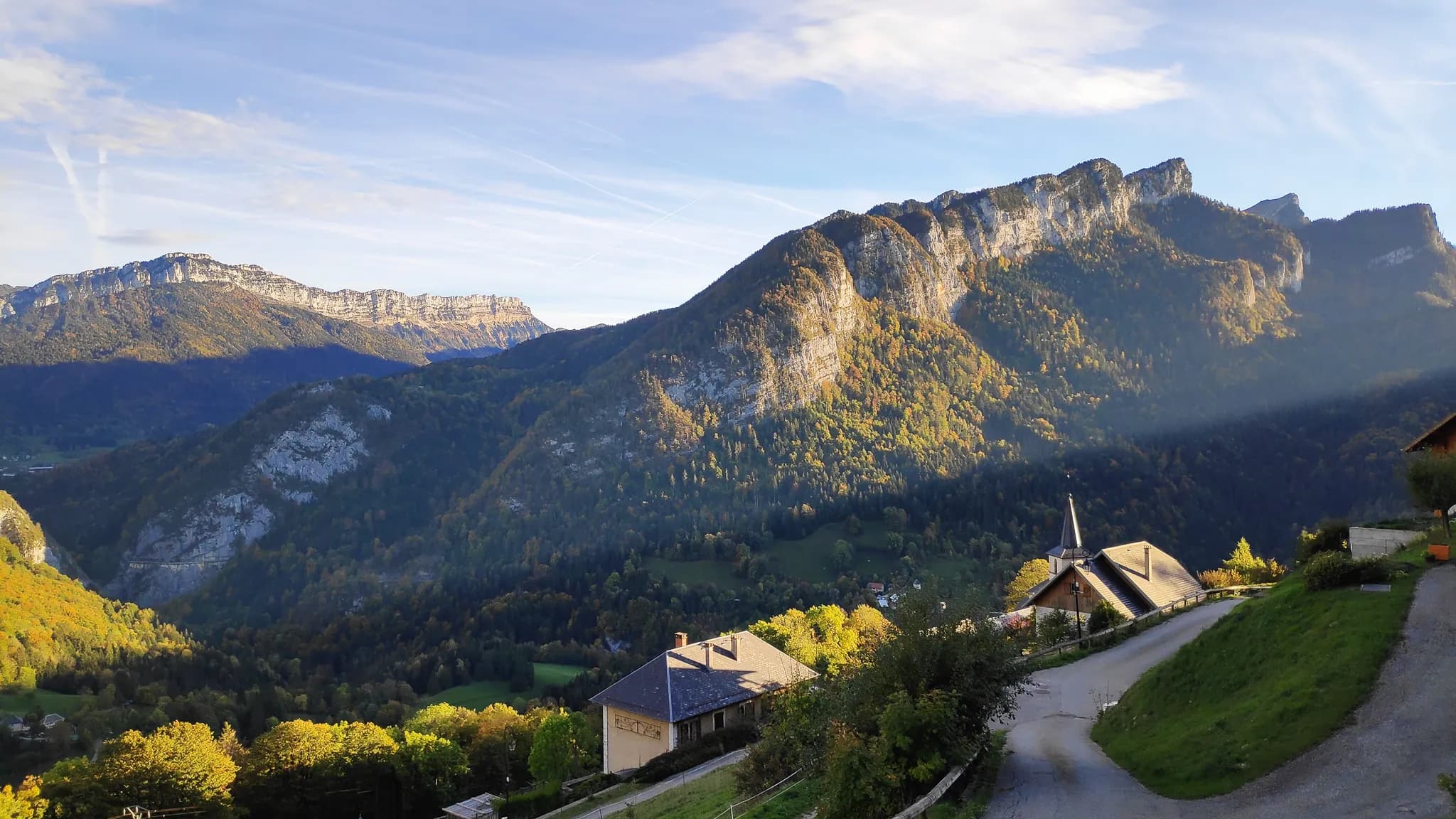 Un paysage montagneux avec des sommets escarpés dominés par des falaises. Au premier plan, on aperçoit plusieurs bâtiments, dont un chalet avec un toit en pente et une église à flèche élancée. La végétation autom