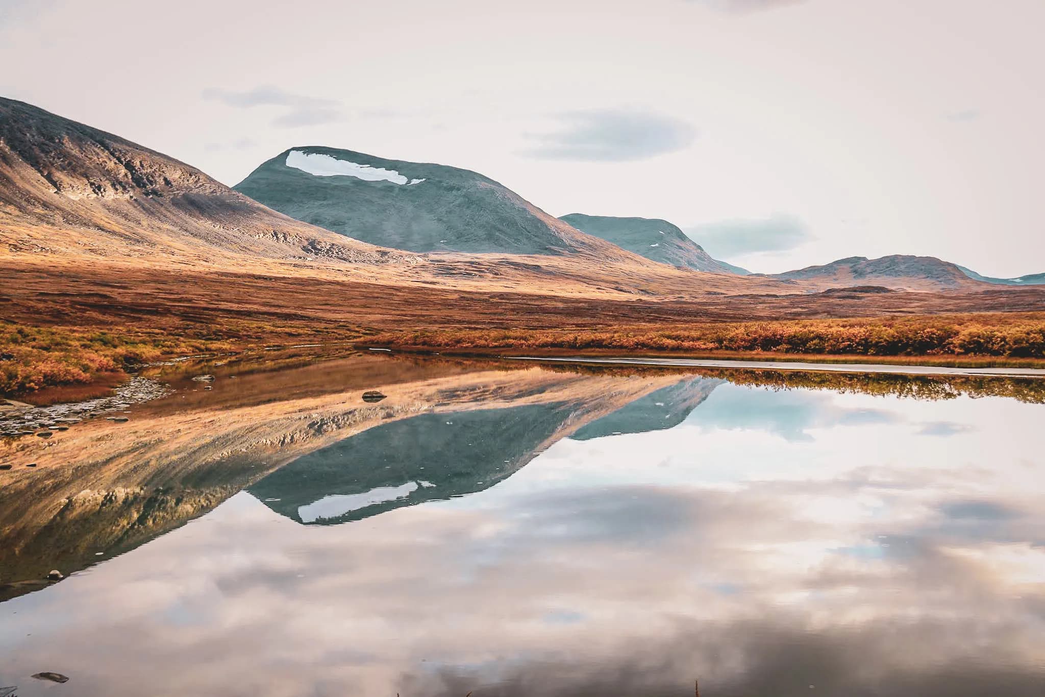 A serene natural landscape reflecting majestic mountains in a calm pond. The hills are lined with golden and orange autumn vegetation, while the peaks still bear a few traces of snow.