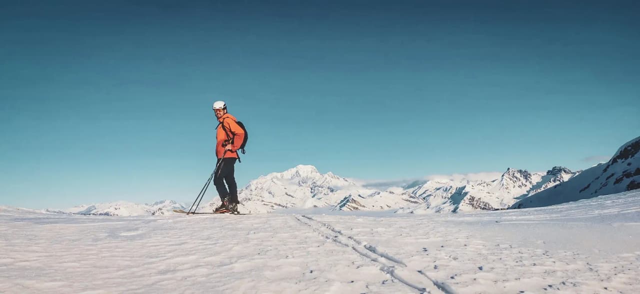 A skier in the mountains, facing a snow-covered landscape and a brilliant blue sky. An off-piste adventure!