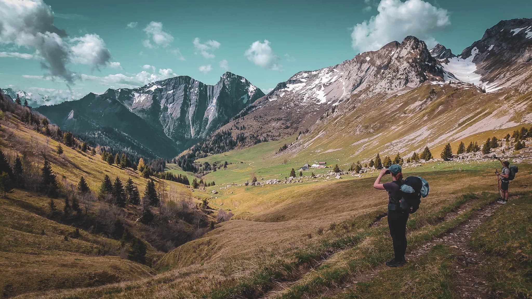 Un paysage de montagne avec des sommets majestueux, certains couverts de neige, et une vaste vallée verdoyante. Au premier plan, des randonneurs avec des sacs à dos admirent la vue. Des conifères parsè