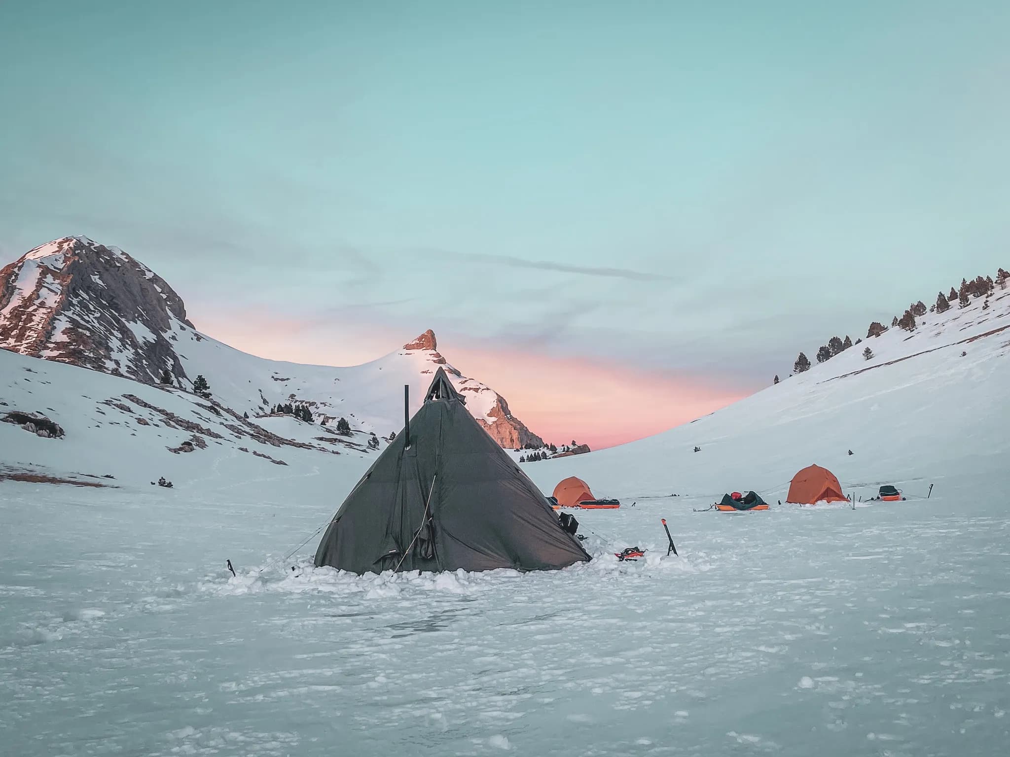 A snow-covered camp with several tents, including a cone-shaped main tent, is in the foreground, while snow-covered mountains rise in the background. The sky is coloured