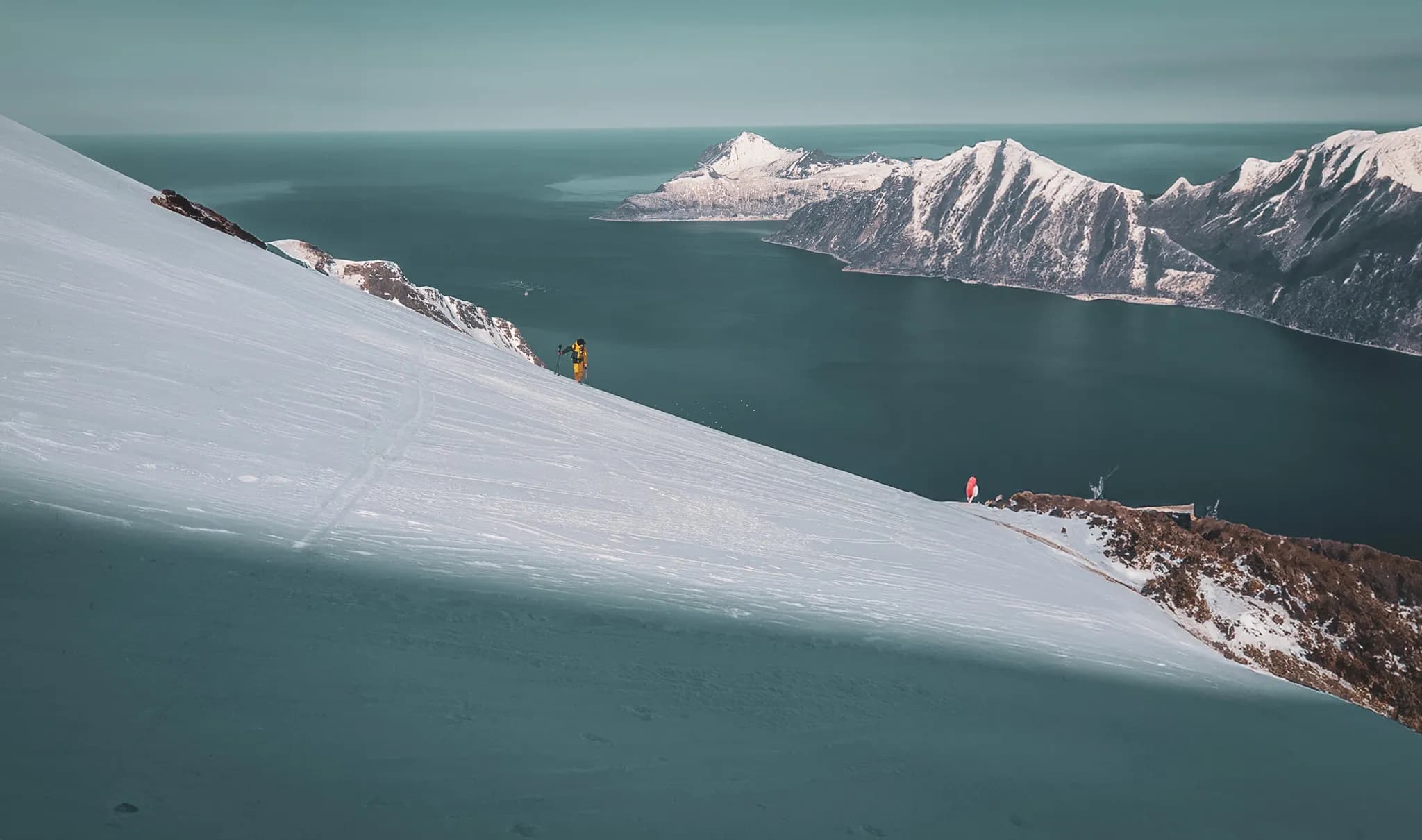 Een besneeuwd berglandschap, met een zachte, besneeuwde helling die naar beneden leidt naar een kalme zee. Op de achtergrond rijzen majestueuze bergen op, sommige met besneeuwde toppen. Een avonturier