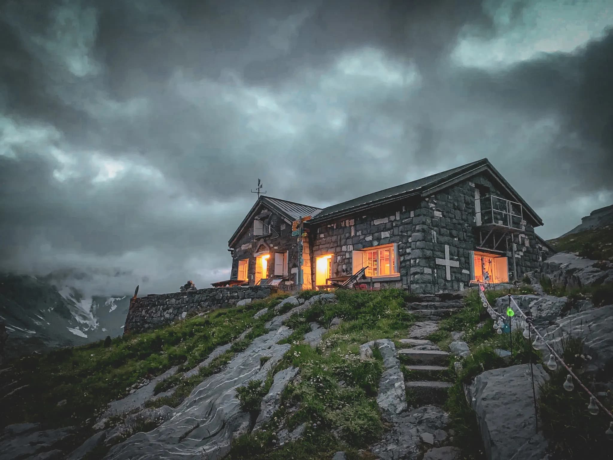 A stone chalet stands on a hill, surrounded by rocks and lush green vegetation. Its windows are illuminated by a warm light, contrasting with the dark, cloudy sky that envelops the whole area.