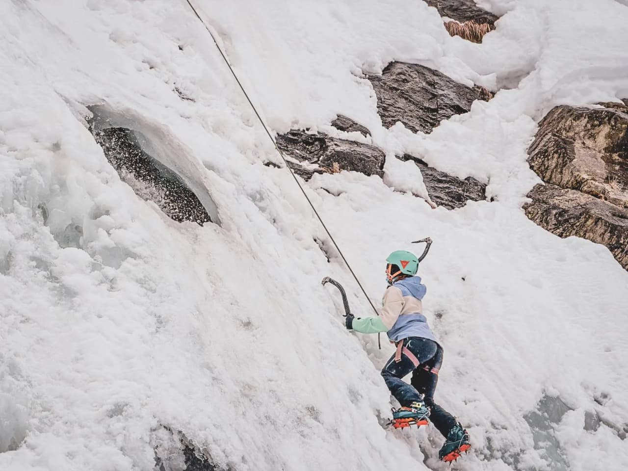 Un alpiniste en pleine ascension sur une cascade de glace, avec des sommets alpins en toile de fond.
