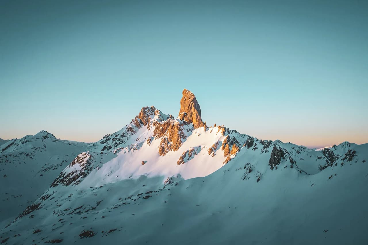 A majestic mountain peak rises above a snow-covered landscape, bathed in the soft light of a sunrise. Snow blankets the slopes, while sheer rock formations emerge, creating the most breathtaking scenery in the world.