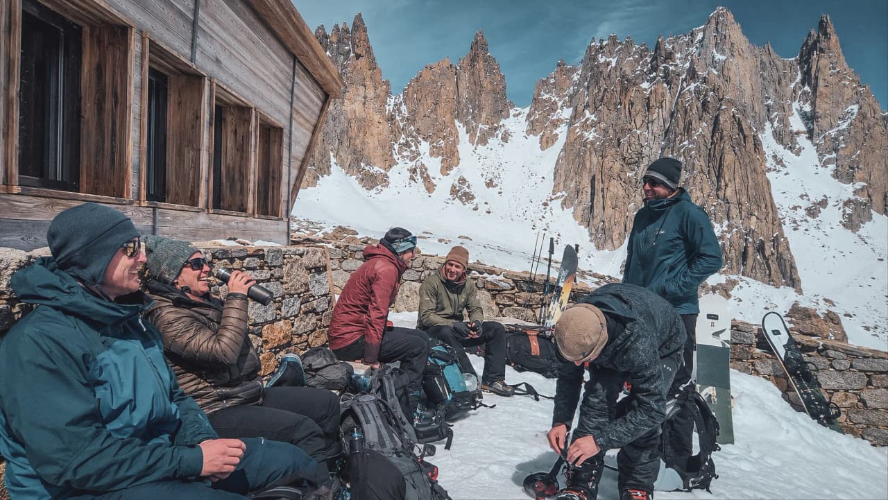 Groupe de snowboarders se reposant près d'un refuge, avec des montagnes majestueuses en arrière-plan.