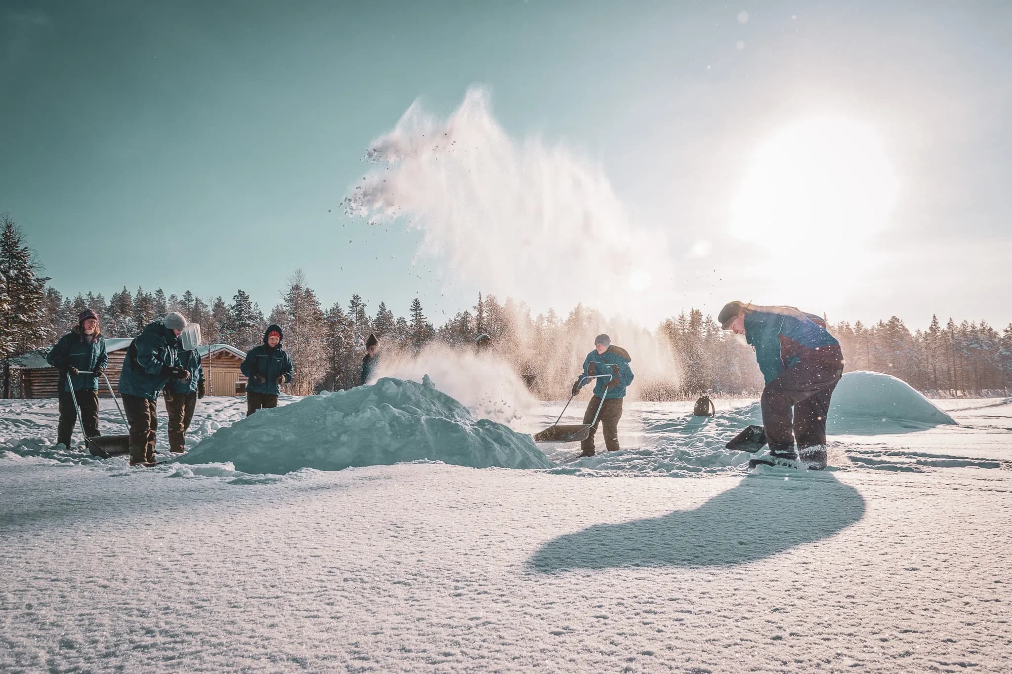 A group of people work in the snow, creating piles and clearing away the flakes in a sunny winter atmosphere. The setting is surrounded by dense trees, while a light cloud of snow