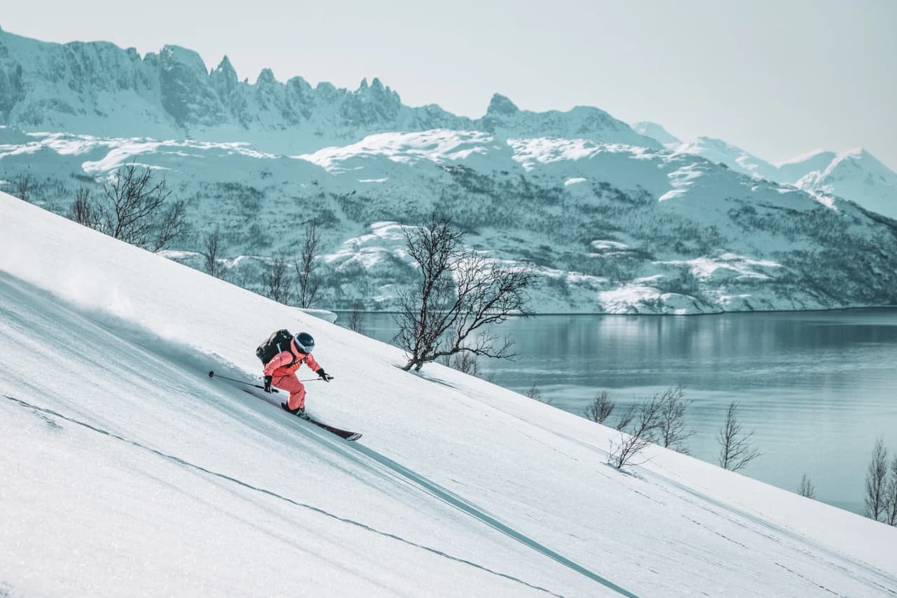 A skier in a pink outfit hurtles down a snowy slope, leaving a trail in the fresh snow. In the background, majestic snow-capped mountains loom under a clear, bright sky. A lake