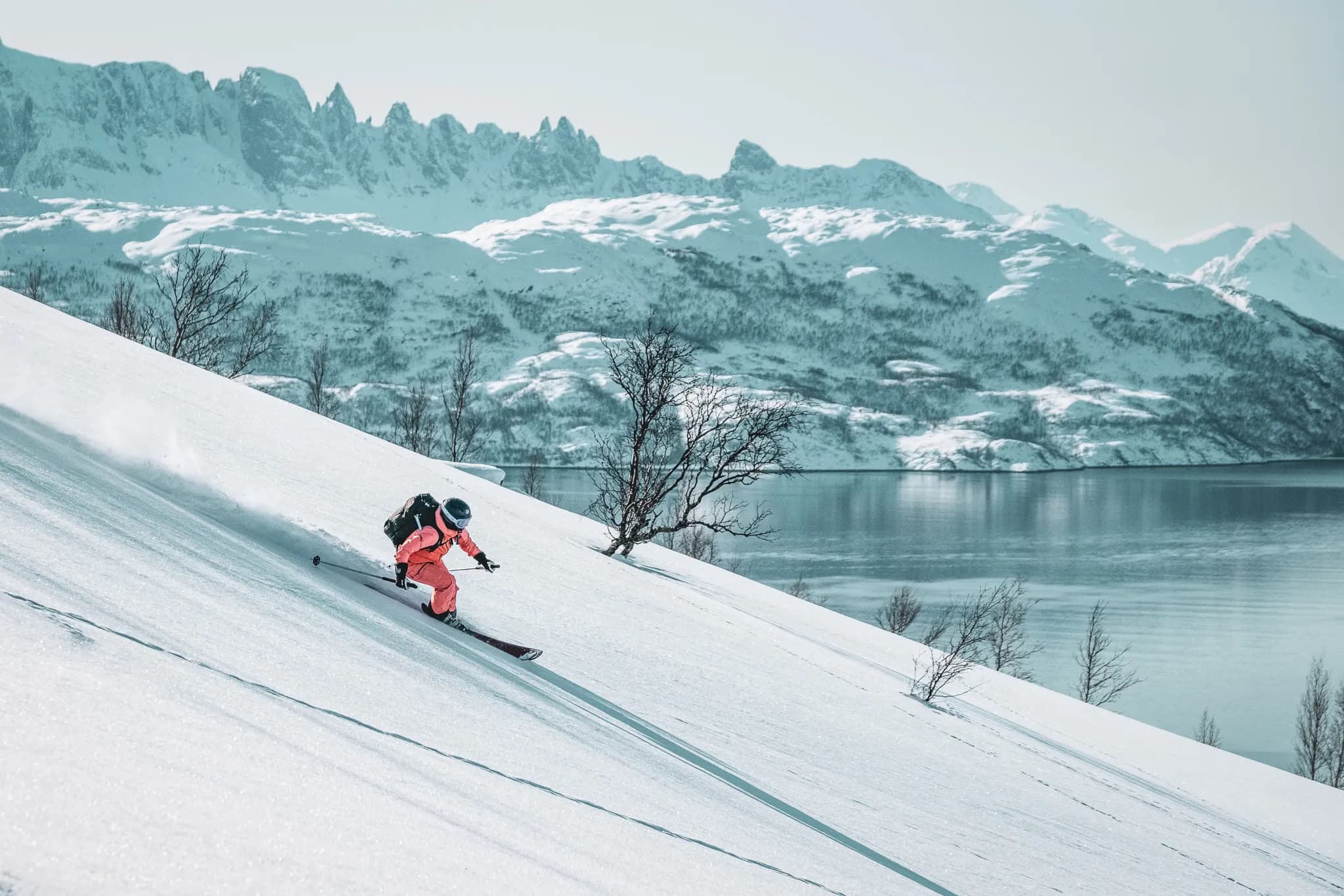 Un skieur en tenue rose dévale une pente neigeuse, laissant une traînée dans la neige fraîche. En arrière-plan, des montagnes majestueuses recouvertes de neige se profilent sous un ciel clair et lumineux. Un lac