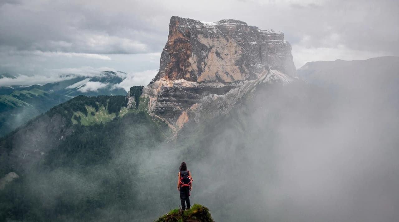Un randonneur contemplant un majestueux sommet dans les nuages des Hauts Plateaux du Vercors.
