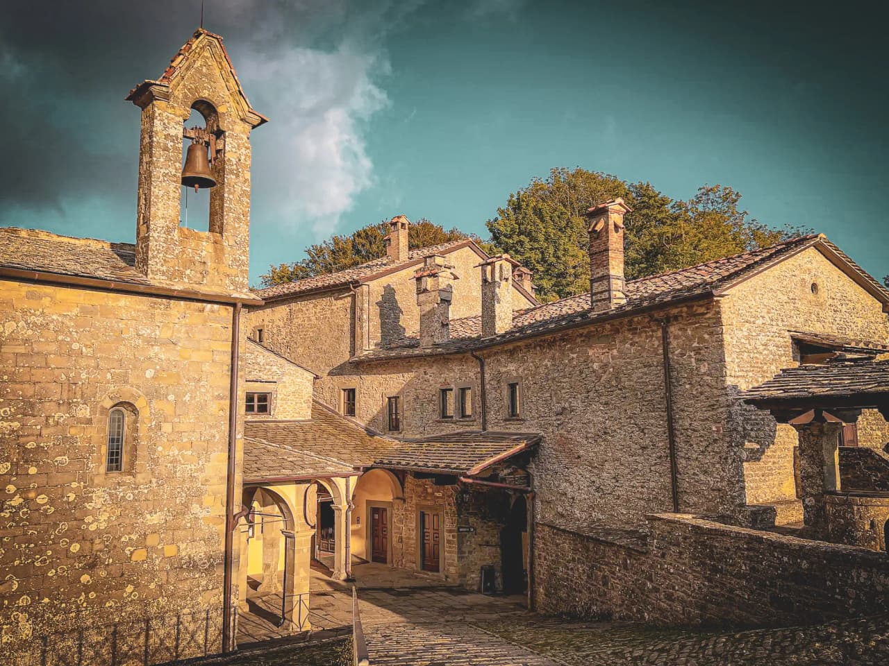 An old building, probably a church or monastery, with a bell visible in a stone bell tower. The stone wall is adorned with lichens, and the roof is tiled.