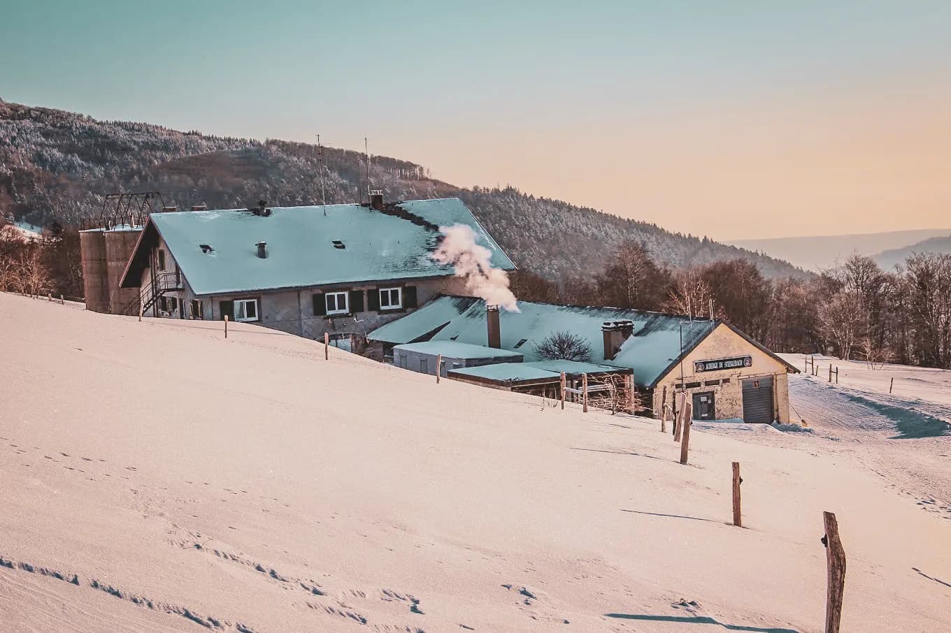 A wooden building covered in snow, set in a wintry landscape. The building's chimney is emitting smoke, indicating a source of heat inside. The surrounding terrain is covered with a thick layer of