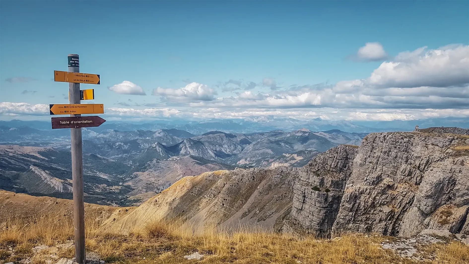 A signpost in a vast and spectacular mountain landscape, with different directions indicated for destinations such as "Sommet des Moulins" and "Table d'orientation". The view captures jagged peaks