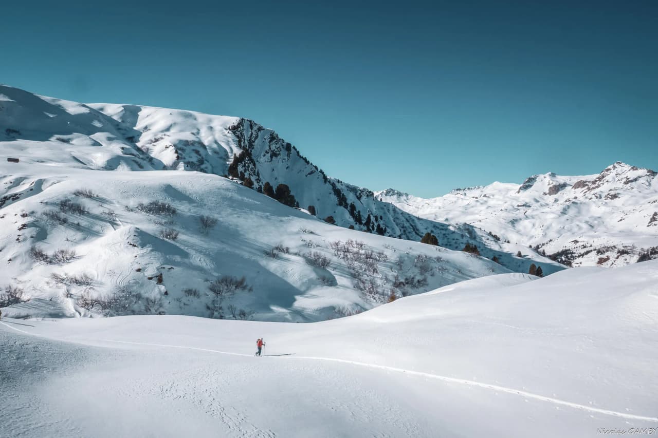 A snowy mountain landscape, with gentle hills covered in a thick layer of snow. In the background, majestic peaks with steep slopes loom under a clear blue sky. Visit