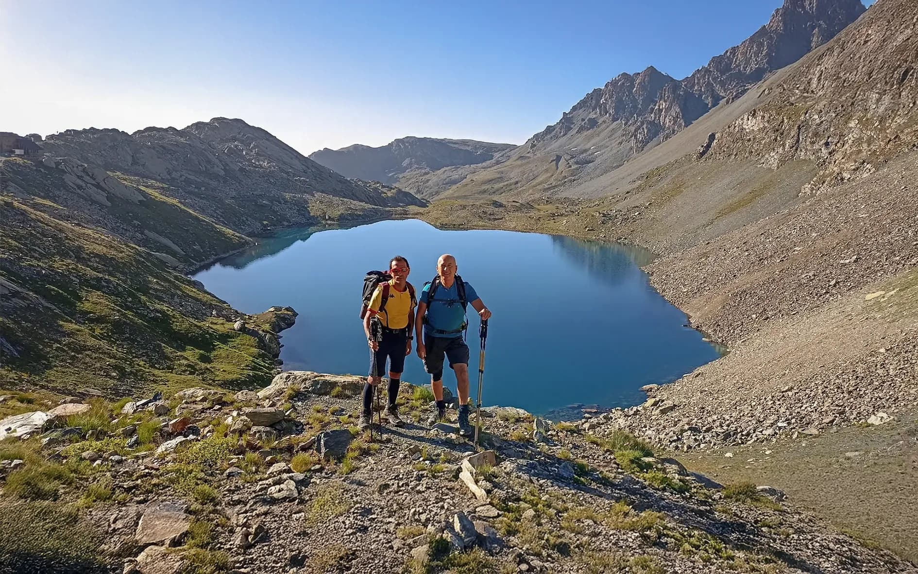Two smiling hikers in front of a turquoise lake, surrounded by majestic mountains.