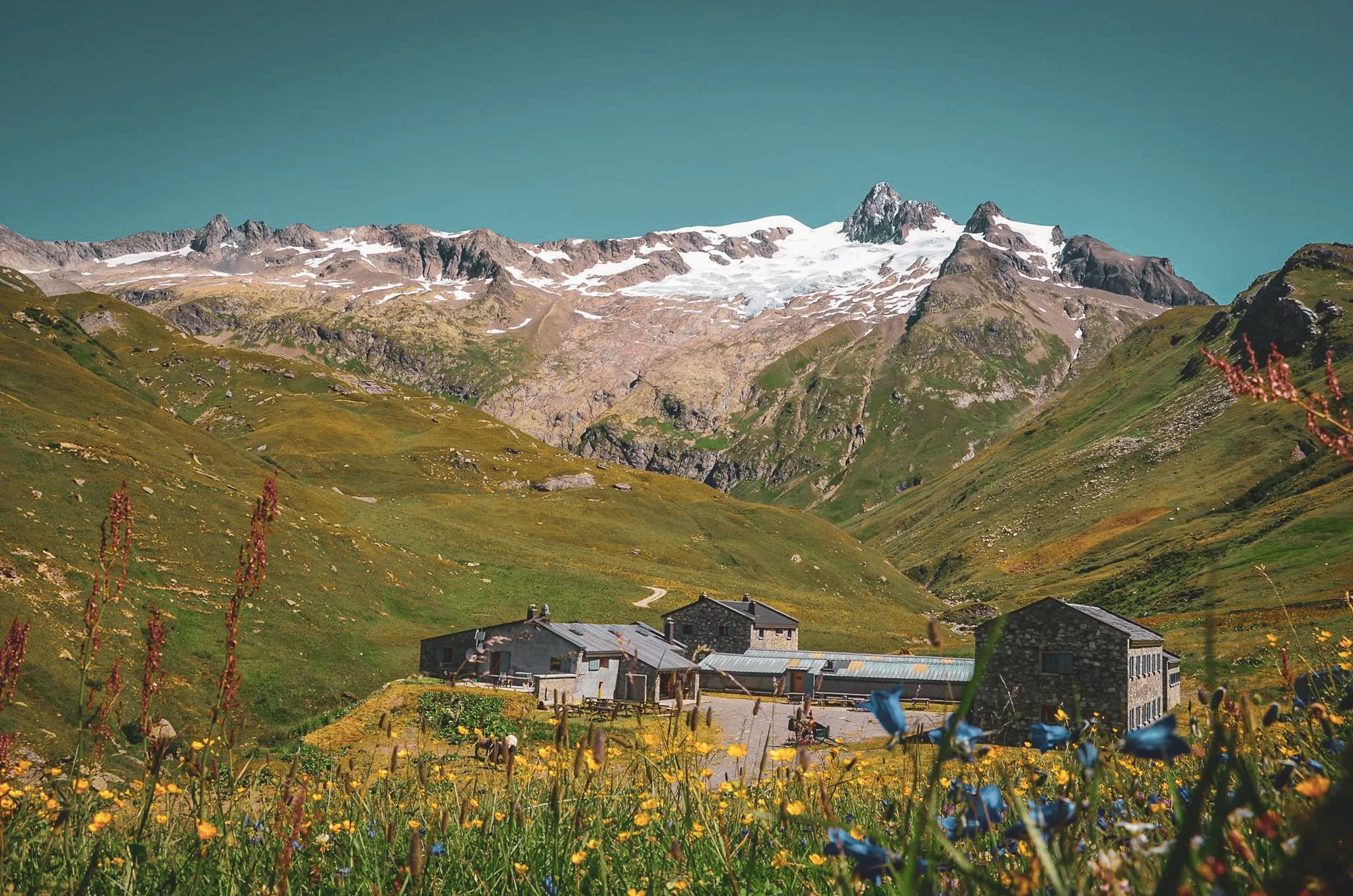 Un paysage alpin majestueux avec des montagnes recouvertes de neige en arrière-plan, où les sommets rocheux se détachent contre un ciel bleu vif. Au premier plan, un bâtiment rustique en pierre se trouve entouré d