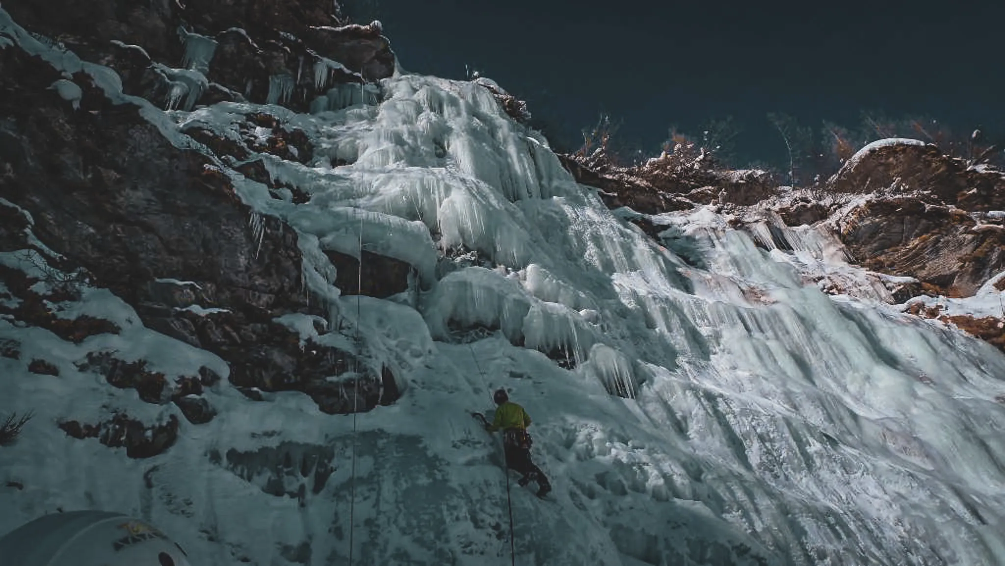 Een bergbeklimmer beklimt een glinsterende ijsval in de Italiaanse Alpen, onder een donkere hemel.