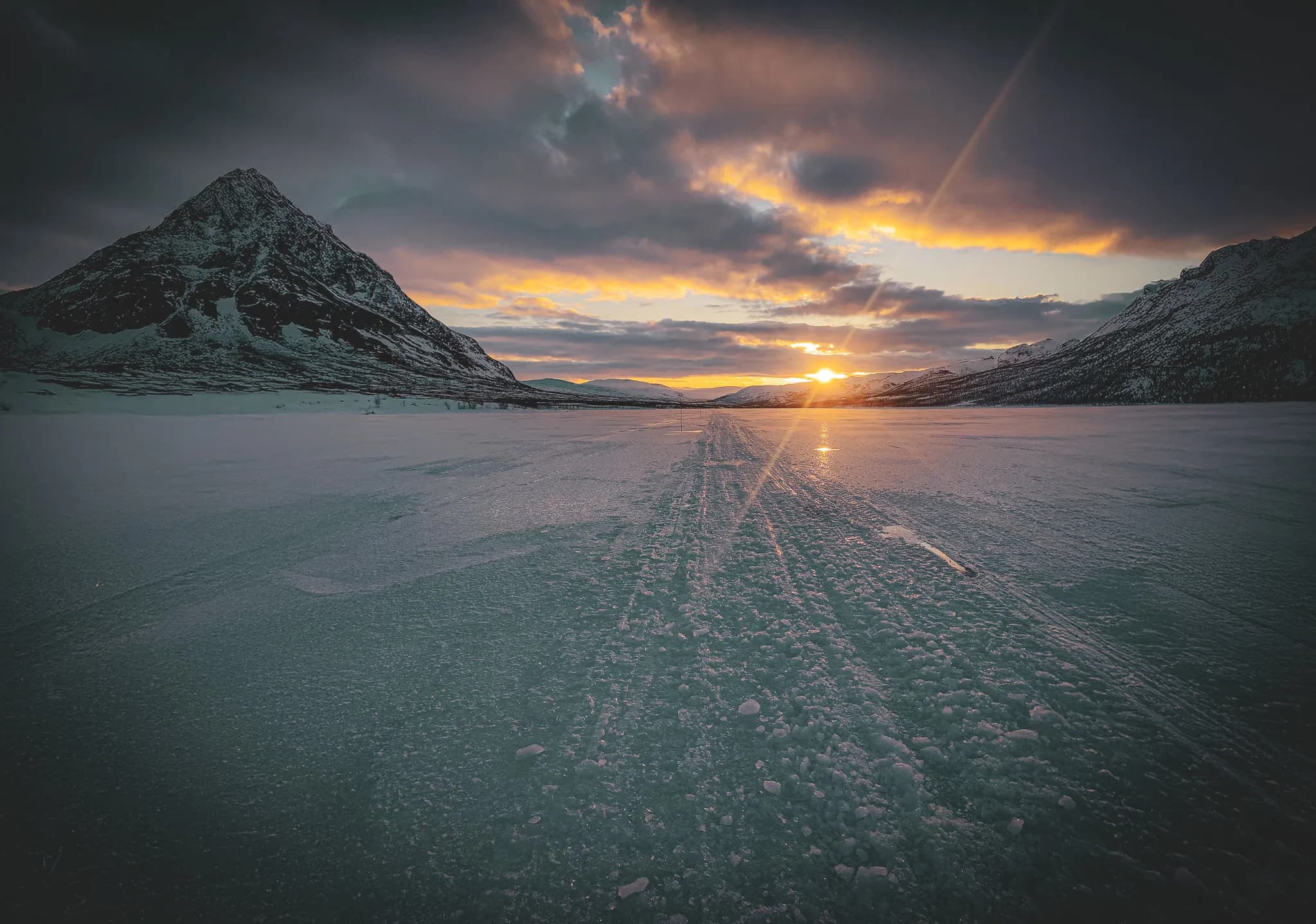 A mountainous landscape unfolds, with snow-capped peaks under a cloudy sky. A frozen lake stretches as far as the eye can see, with glide marks visible on the icy surface.