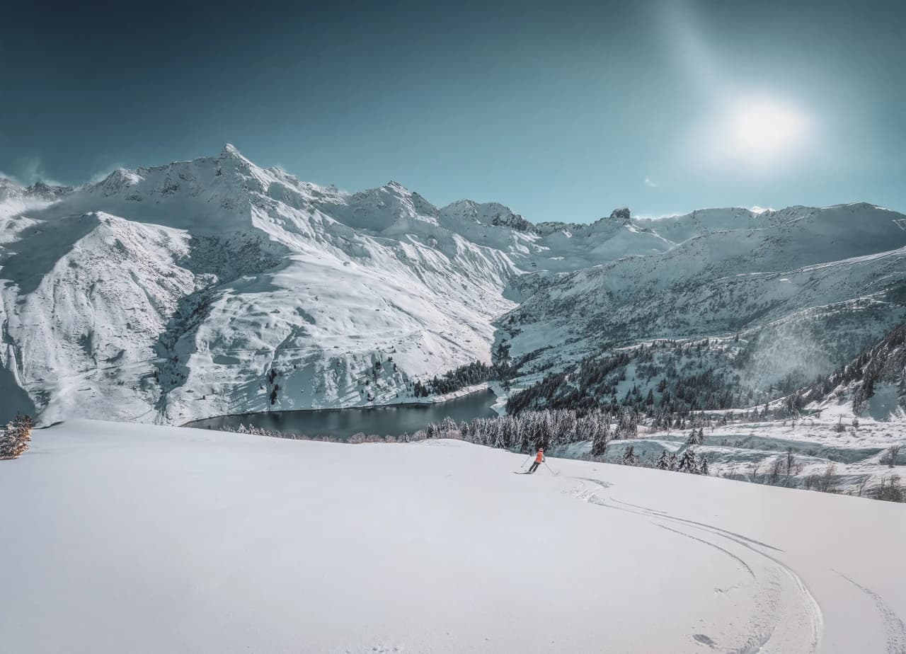 An Alpine landscape in winter, with snow-capped mountains towering majestically beneath a clear blue sky. In the foreground, an expanse of untouched snow bears witness to recent snowfalls. A sk