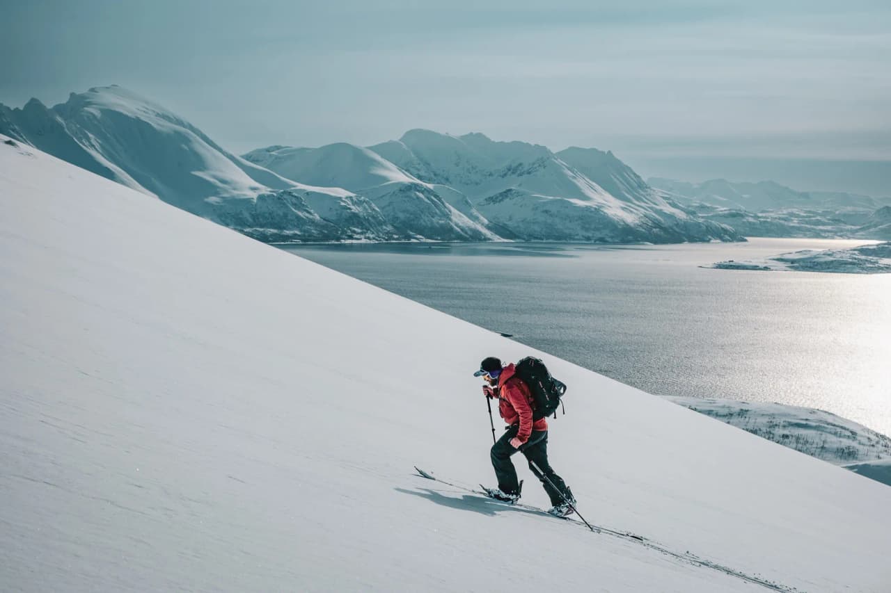 A skier in full effort makes his way up a snow-covered slope, equipped with a rucksack and ski poles. In the background, a vast expanse of snow-covered mountains rises majestically.