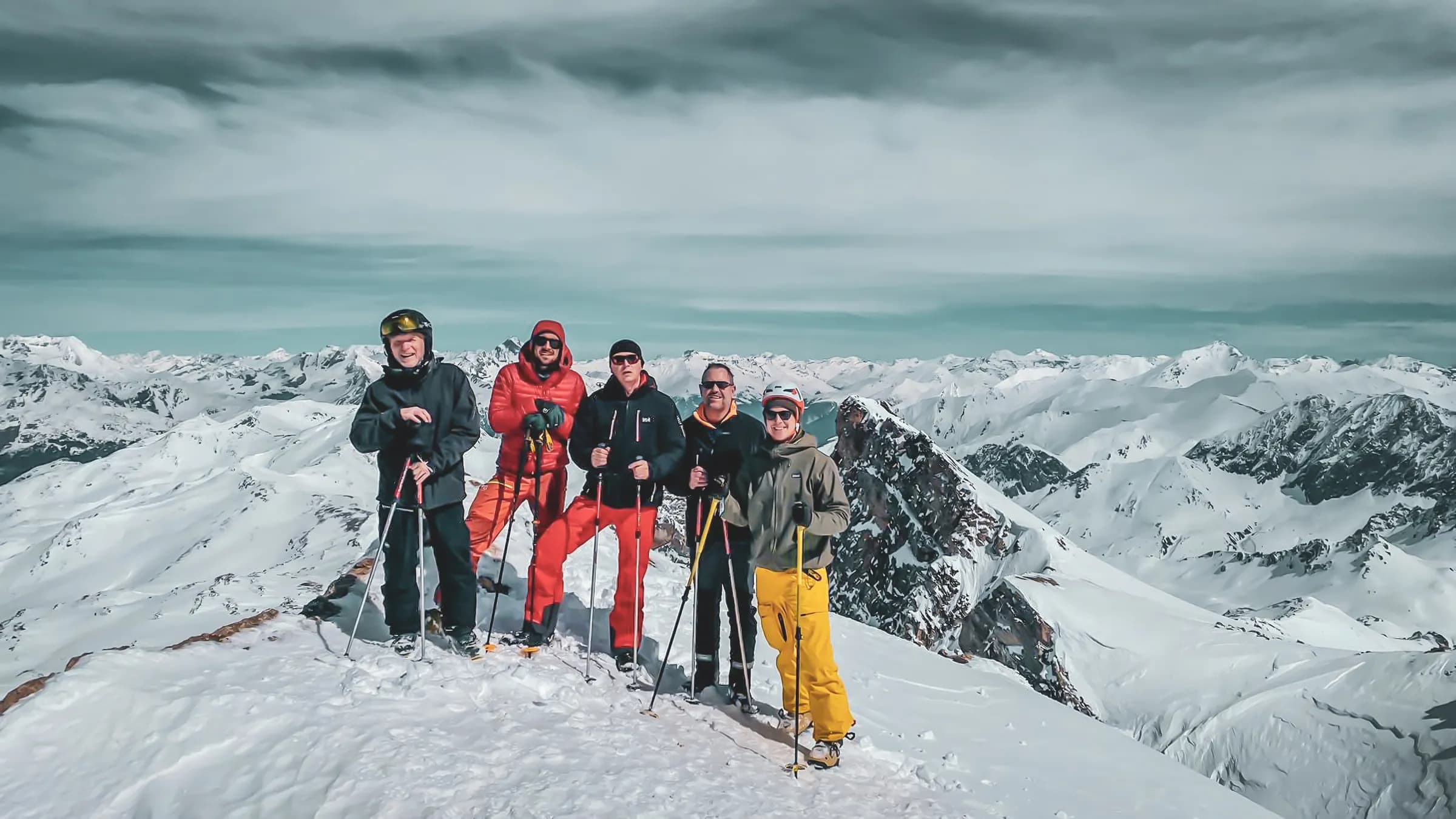 Un groupe de cinq personnes se tient sur un sommet enneigé, entouré de montagnes majestueuses recouvertes de neige. Chacune des personnes porte des vêtements d'hiver colorés et tient des bâtons de ski. L'un