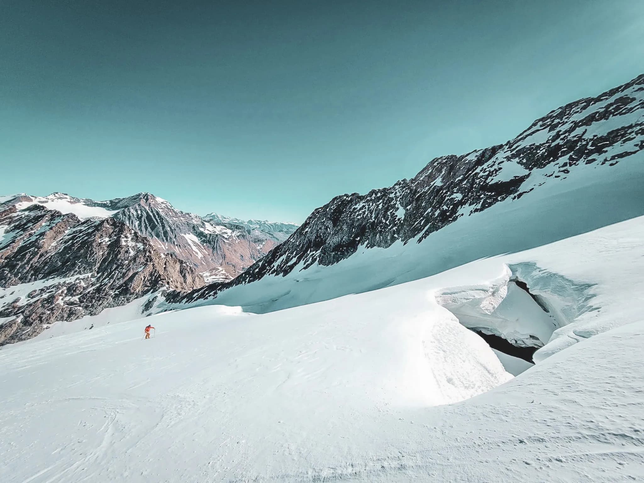 A snow-covered high mountain landscape, with majestic rocky ridges in the background. Snow covers the ground, creating a striking contrast with the clear blue sky. A lone skier, dressed