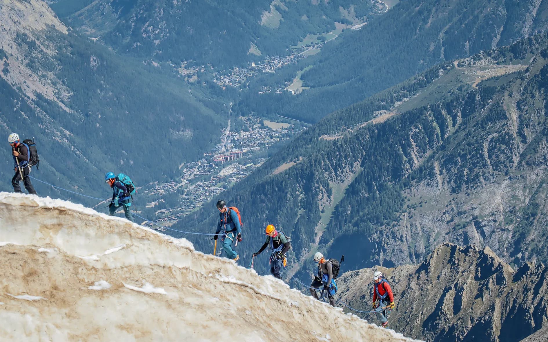 A group of climbers scale an icy slope, overlooking a green valley and a village below. The climbers are wearing helmets and climbing gear, a reminder of their commitment to