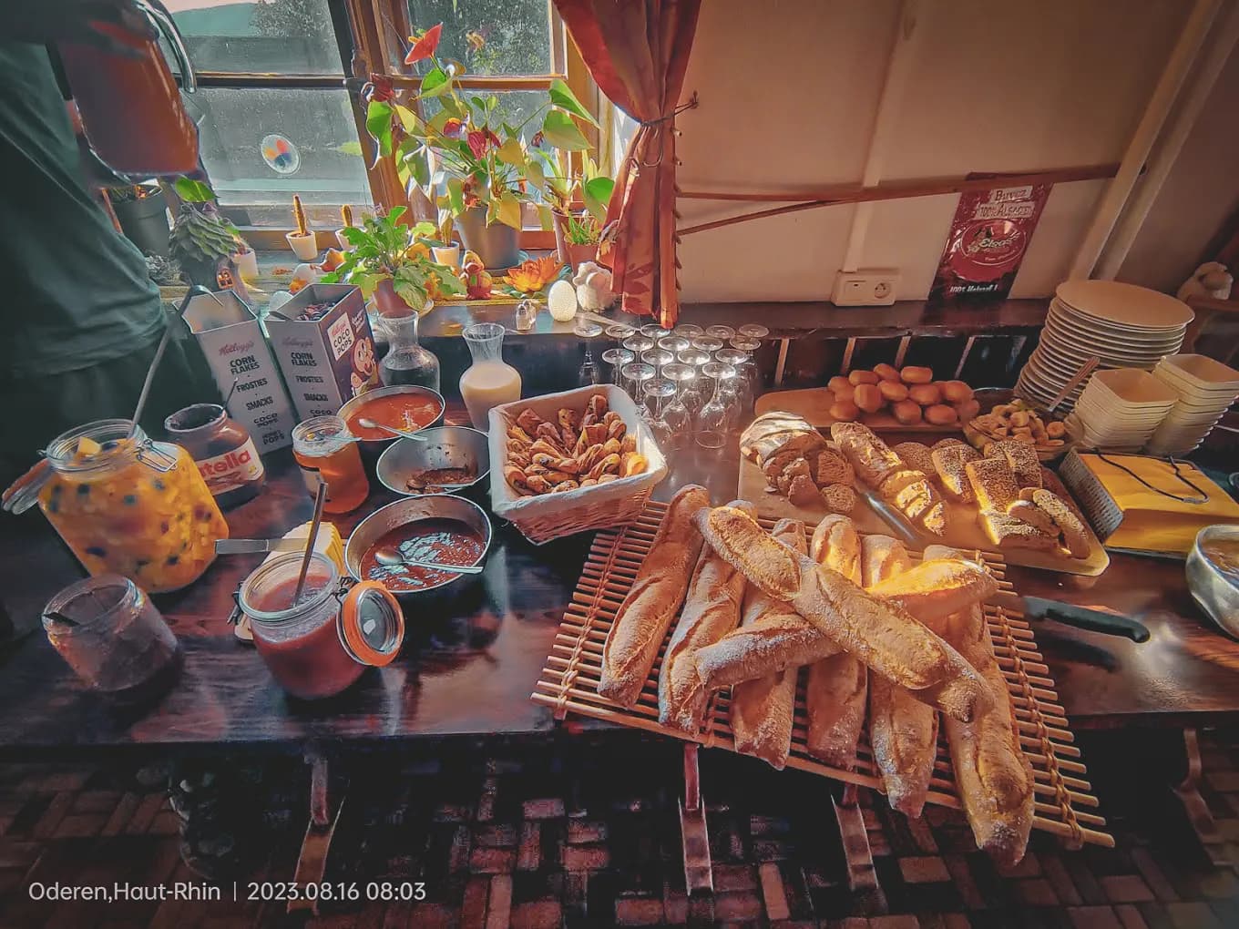 A well-stocked breakfast buffet is laid out on a wooden table. Several types of bread, including baguettes and rolls, are attractively arranged. Next to it are containers containing jams, yoghurt, yoghurts