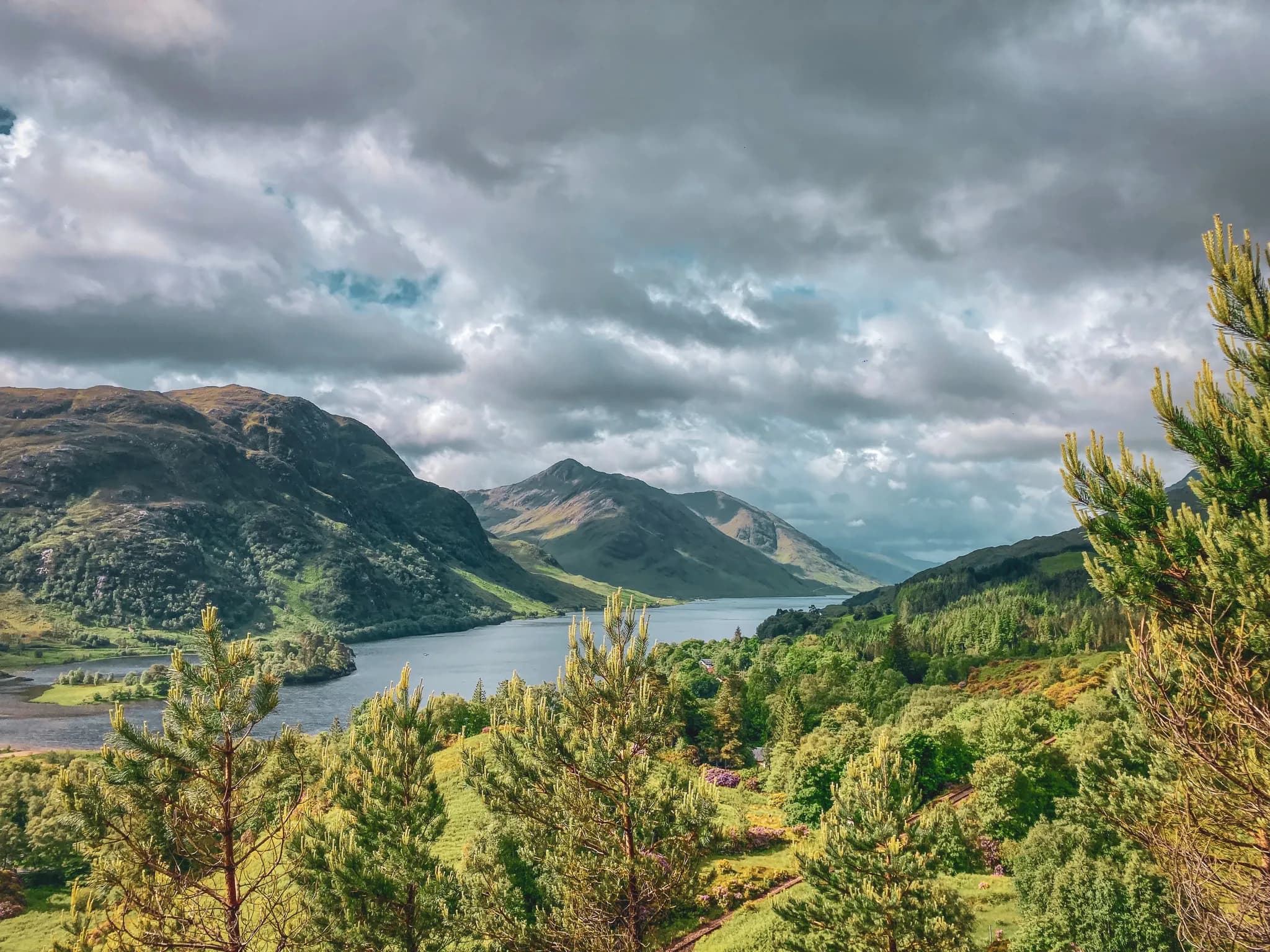 Panoramisch uitzicht op de Schotse Hooglanden, met een sprankelend meer en groene bergen onder een bewolkte hemel.