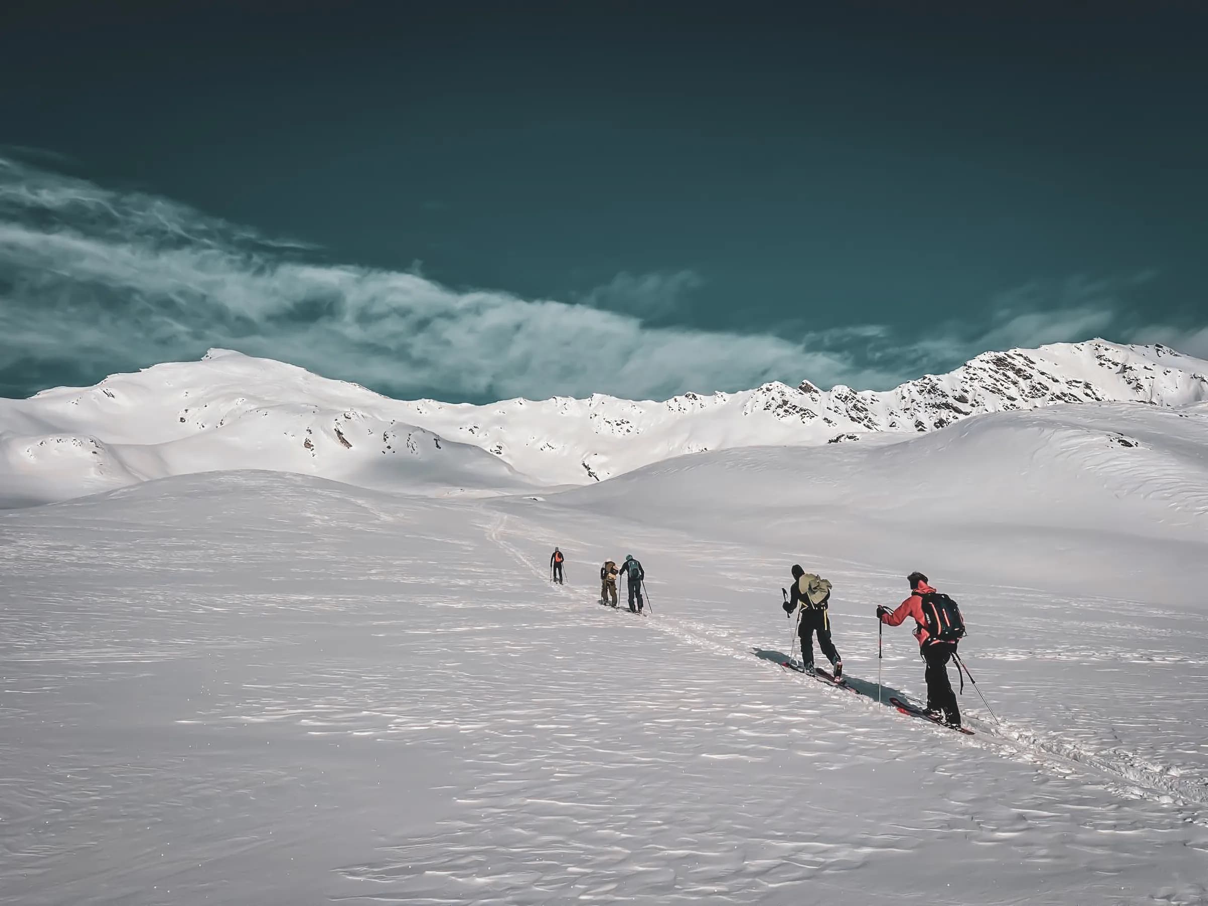 A group of skiers climbing a snowy slope, with snow-capped mountains in the background. The sky is blue with light clouds, adding a peaceful atmosphere to the scene. The skiers